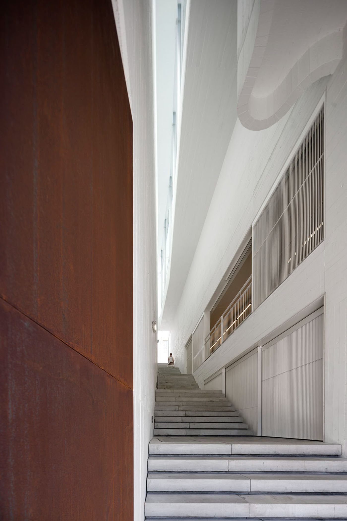 Abrantes Municipal Market by ARX Portugal — A linear staircase ascends through a narrow passage between a textured rust-colored corten steel wall on the left and white rendered surfaces on the right.