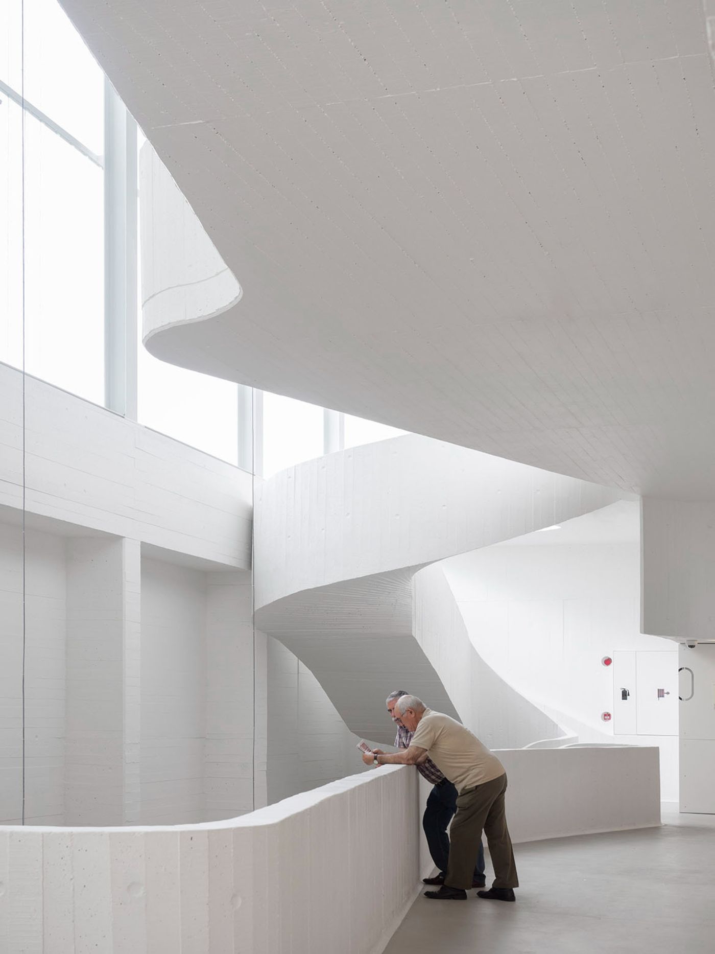 Abrantes Municipal Market by ARX Portugal — An elderly man in a beige cardigan leans against a curved concrete wall in the spiraling interior of the Abrantes Municipal Market.
