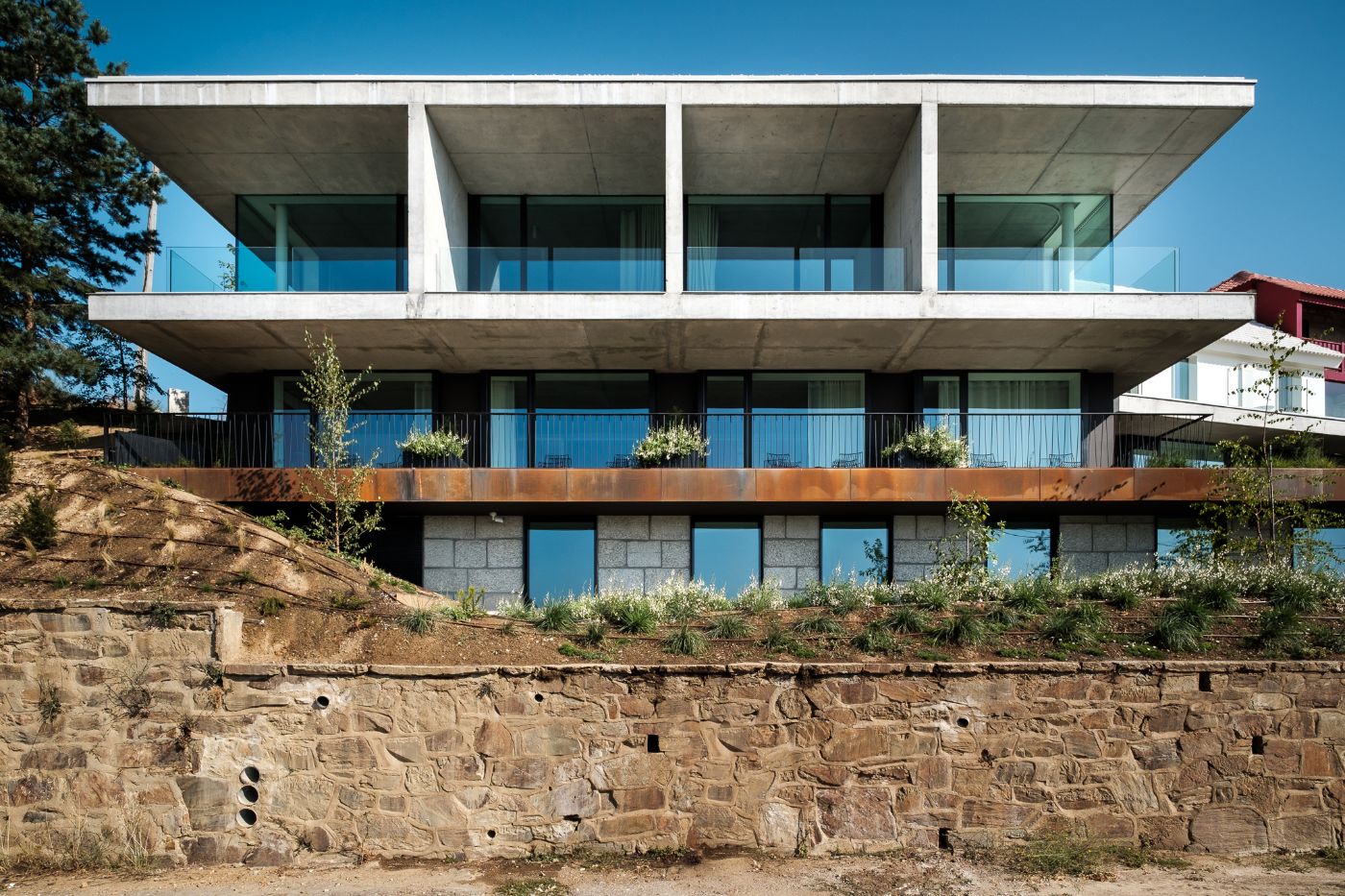 Casa de São Lourenço by P-06 Atelier and Site Specific by P-06, Site Specific, Rogério de Azevedo — A three-story modernist hotel building cantilevers dramatically over a stacked-stone retaining wall, its structural logic exposed through board-formed...