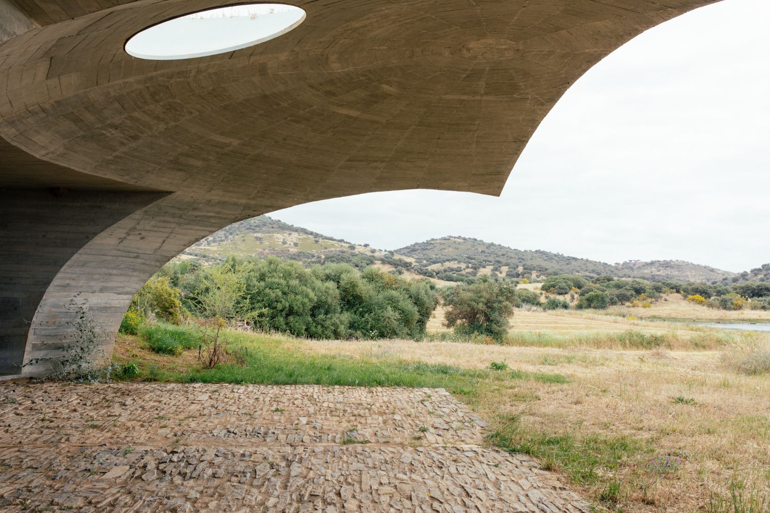 Casa na Terra captured by Thisispaper Studio — Viewed from beneath a monolithic concrete cantilever, the structure frames a rural Alentejo landscape of rolling hills and scattered olive trees.