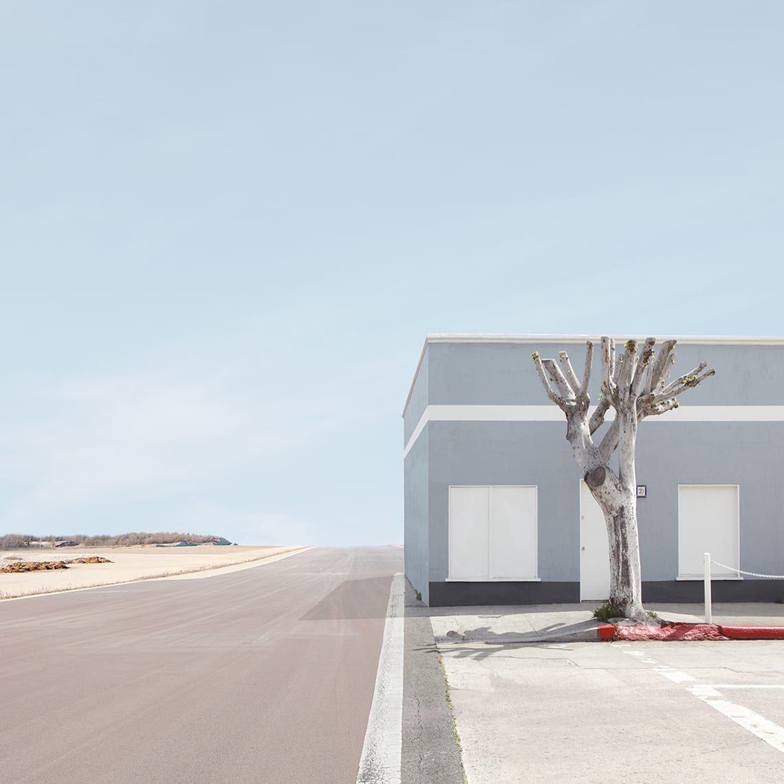 Transition by Lauren Marsolier — A solitary commercial structure punctuates an expansive agricultural landscape, its facade divided by horizontal bands of grey render and cream-colored panels.