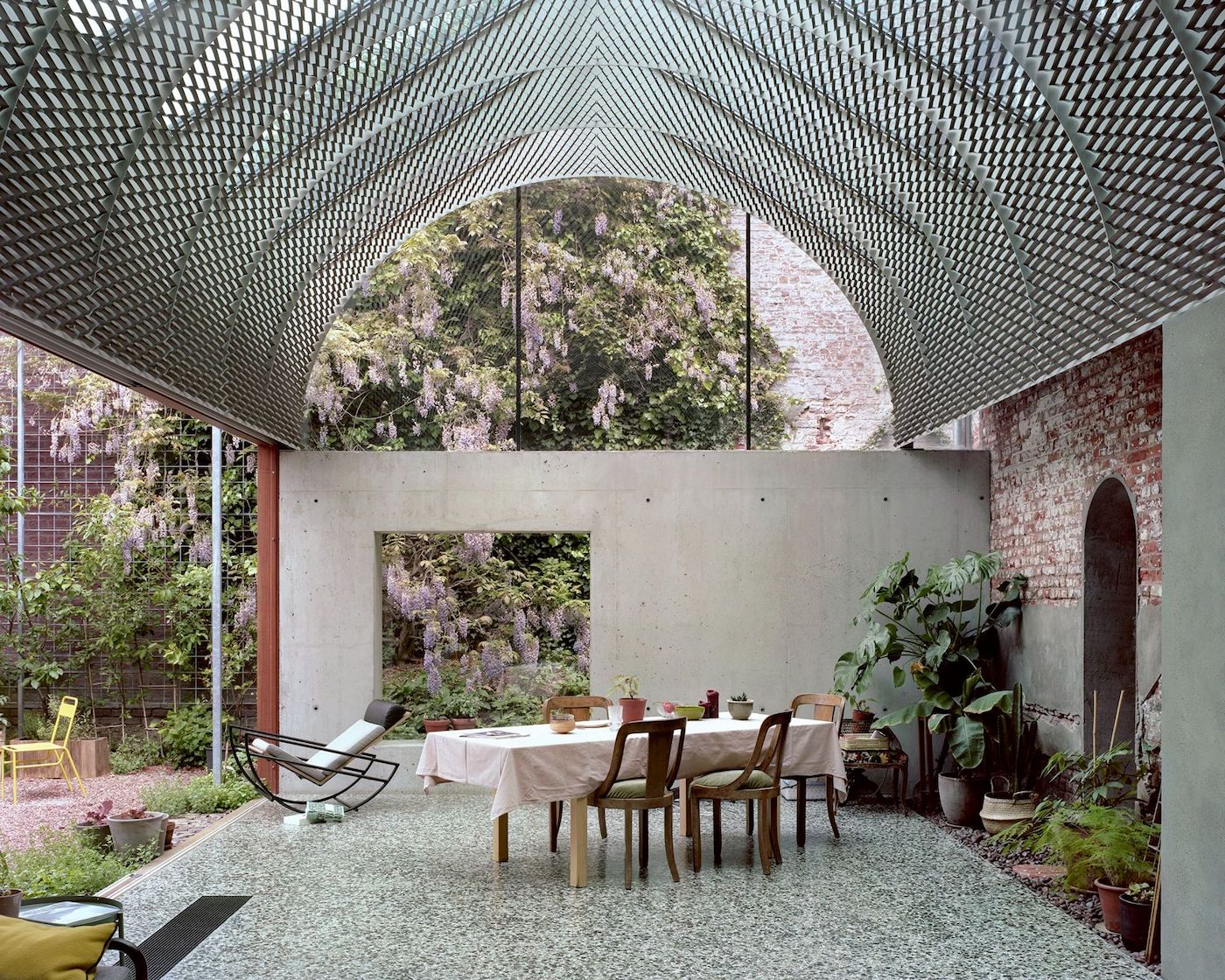 ‘Room in the City’ by 51N4E — A concrete-walled interior space anchored by a round dining table beneath a perforated steel mesh canopy that casts geometric shadow patterns across the...