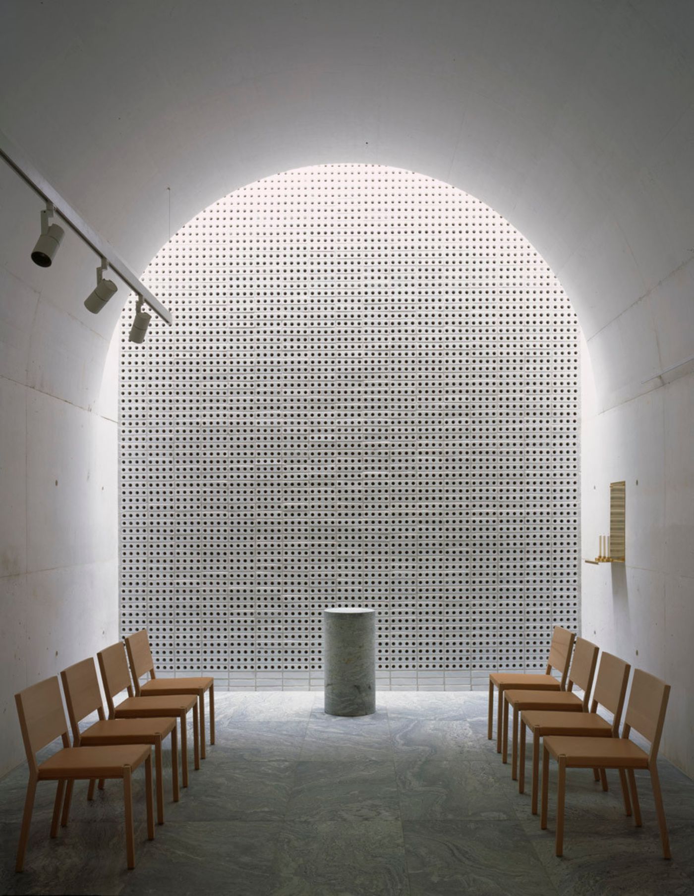 The New Crematorium by Johan Celsing Arkitektkontor — A chapel interior defined by a monumental perforated brick wall with regular grid pattern and arched top, backlit to create luminous depth.