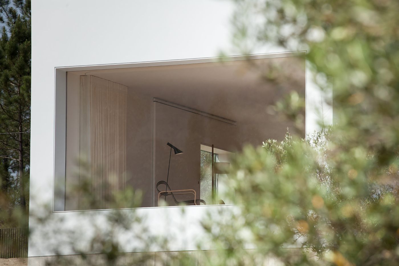 Fonte Boa House by João Mendes Ribeiro — A horizontal strip window frames the rendered white upper volume of Fonte Boa House, its clean facade punctuated by a single black task lamp visible within.