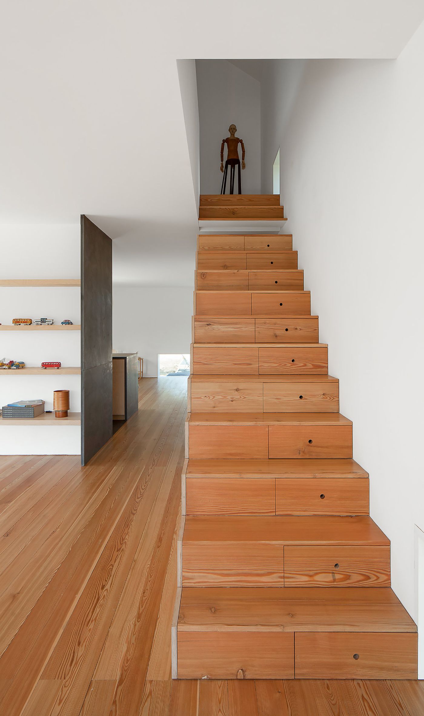 Fonte Boa House by João Mendes Ribeiro — A wooden staircase with integrated storage drawers ascends toward a figure silhouetted against white walls.
