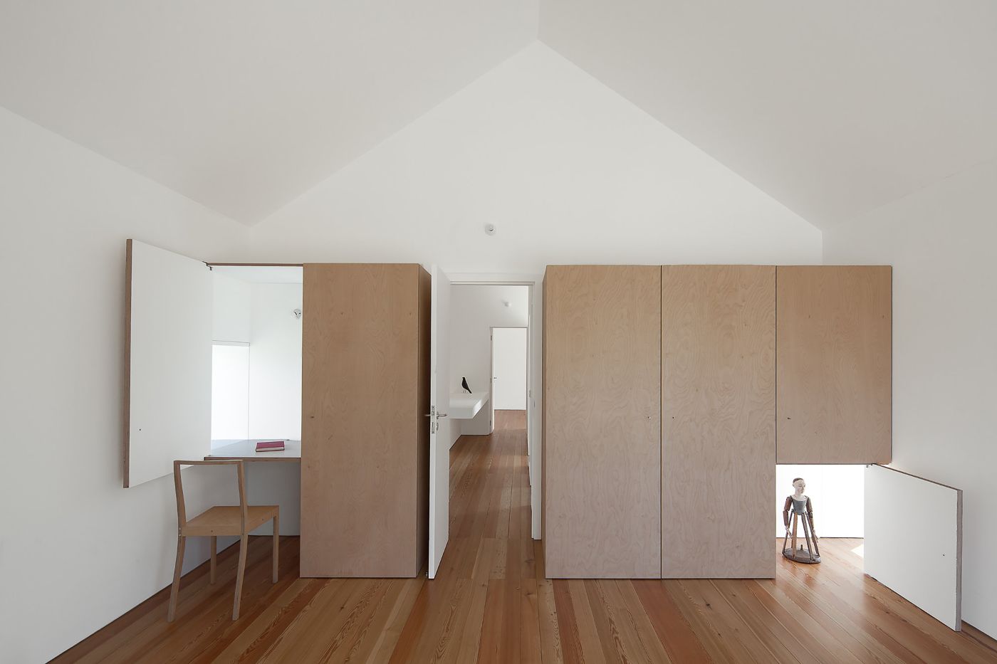 Fonte Boa House by João Mendes Ribeiro — Interior view of a loft-like attic space beneath a pitched roof, furnished with modular plywood partitions and built-in cabinetry in natural wood tones.