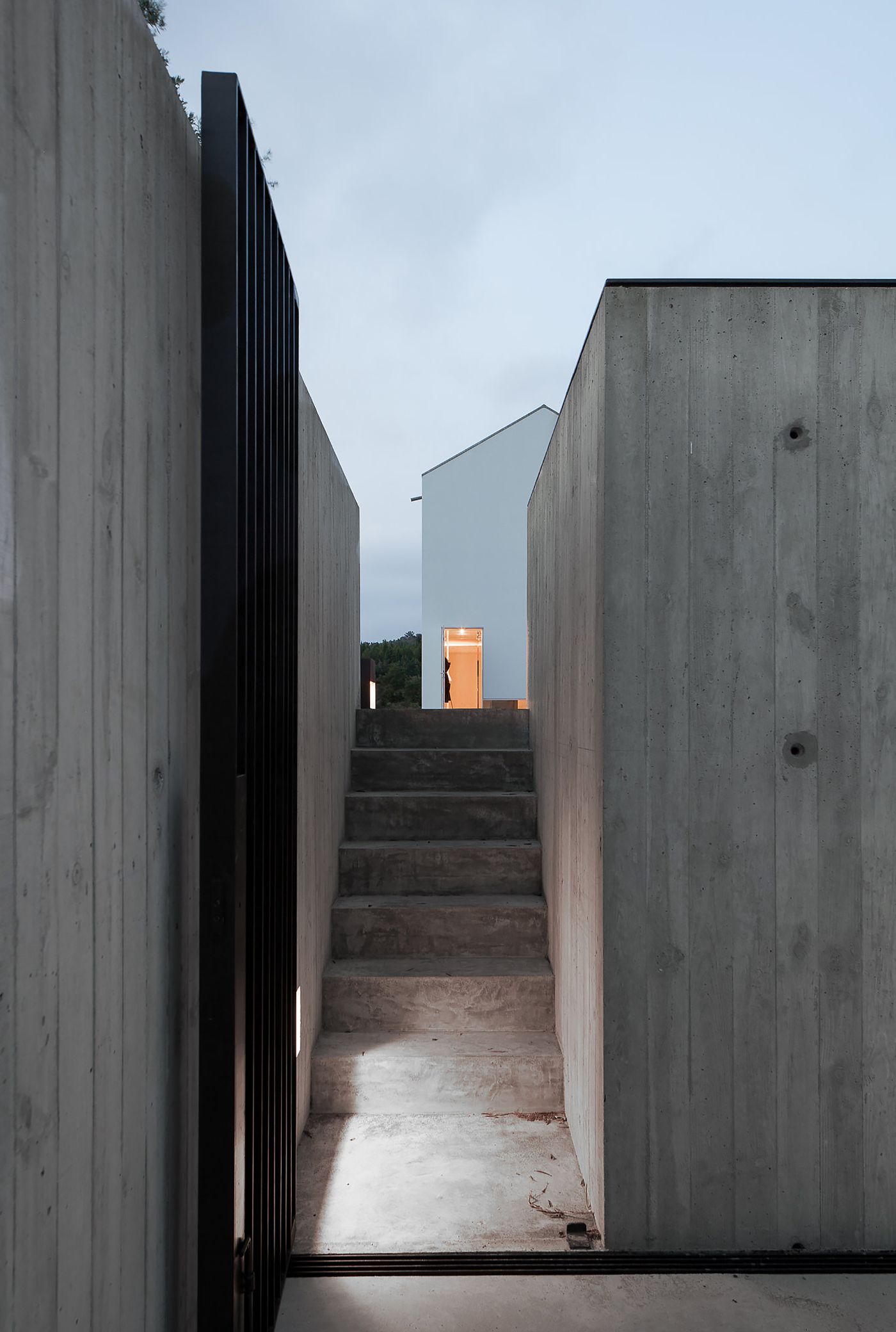 Fonte Boa House by João Mendes Ribeiro — A narrow concrete passage frames the view toward the house's upper volume, rendered white and set against a darkening sky.