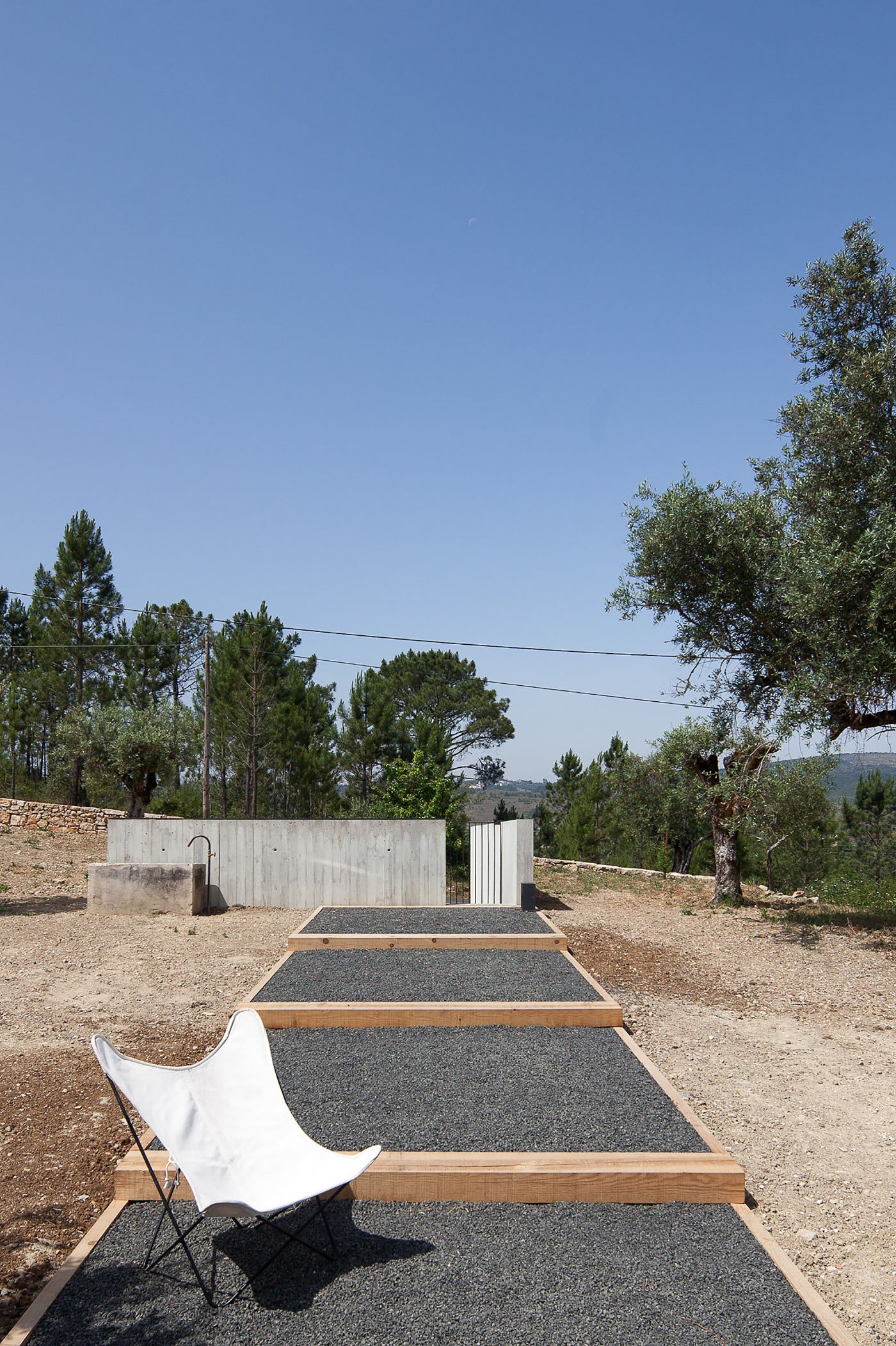 Fonte Boa House by João Mendes Ribeiro — A white butterfly chair sits in sharp shadow on the gravel forecourt, positioned before a minimalist concrete structure with timber-framed edges.