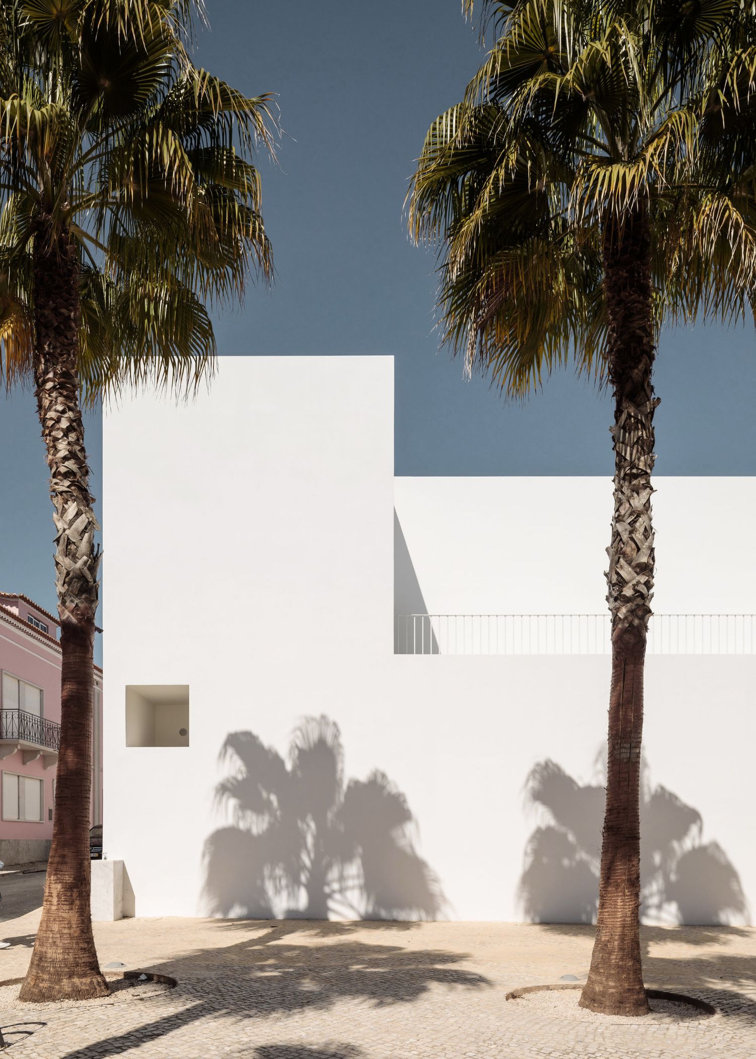Grândola’s Library and Municipal Archive by Matos Gameiro Arquitectos + Pedro Domingos Arquitectos — The rendered white facade of Grândola's Library presents a study in geometric shadow play, with palm fronds casting sharp linear patterns across the...