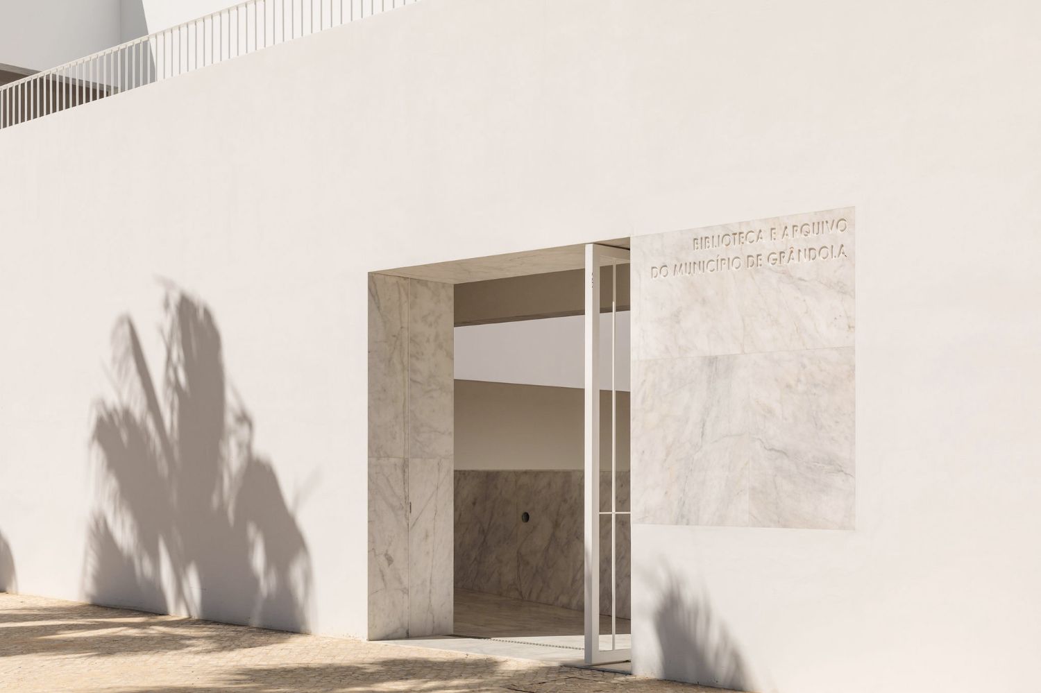 Grândola’s Library and Municipal Archive by Matos Gameiro Arquitectos + Pedro Domingos Arquitectos — The library entrance presents itself as a recessed portal within the rendered white facade, framed by marble cladding on both outer walls.