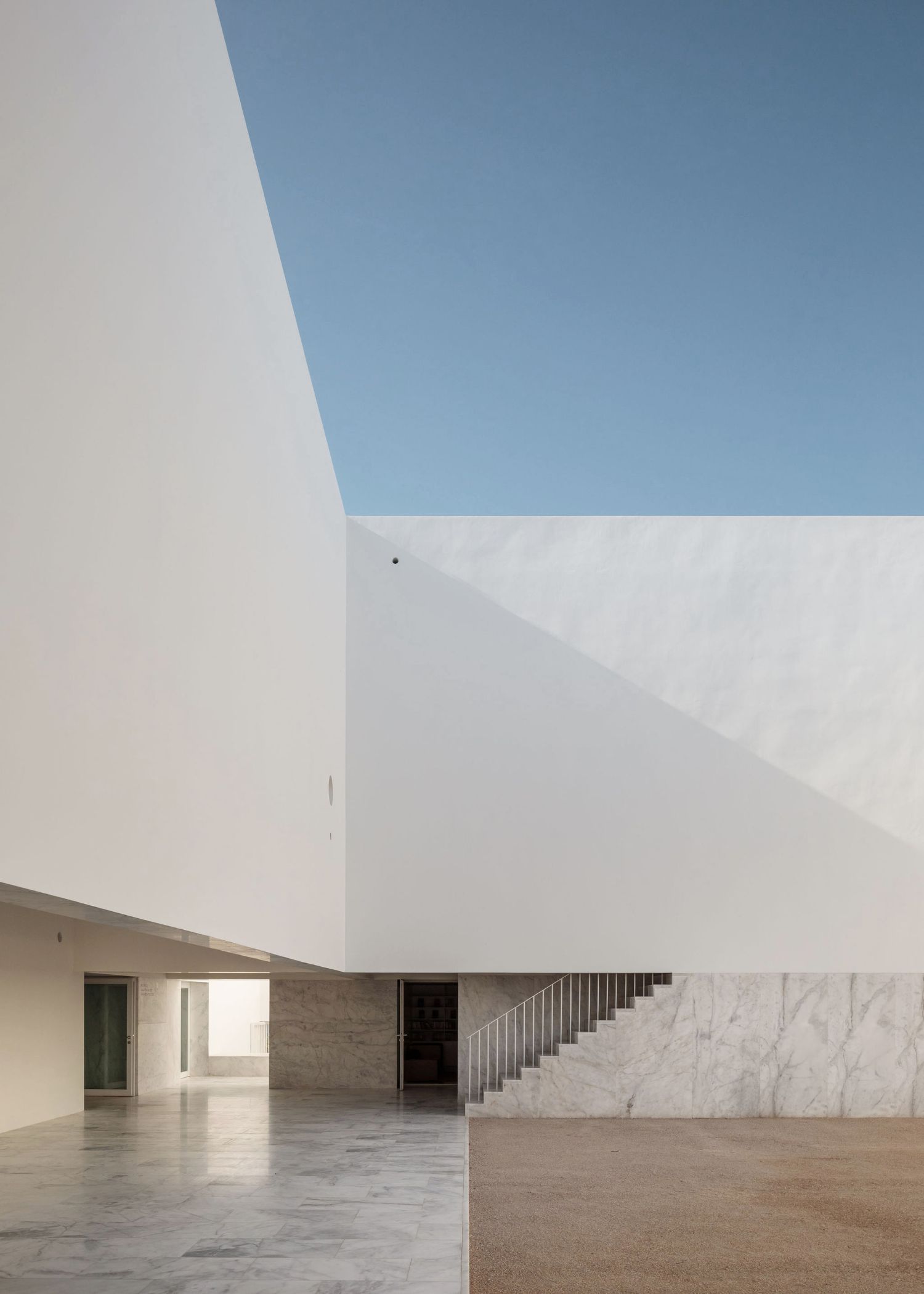 Grândola’s Library and Municipal Archive by Matos Gameiro Arquitectos + Pedro Domingos Arquitectos — A rendered white facade rises above a plaza flooded with sharp shadows cast by unseen palm fronds, creating geometric patterns across the monolithic...