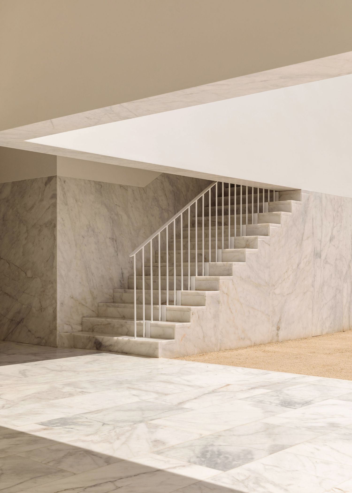 Grândola’s Library and Municipal Archive by Matos Gameiro Arquitectos + Pedro Domingos Arquitectos — A monolithic marble staircase descends into a sunlit plaza, its rendered white walls interrupted by a grid of thin steel railings that cast linear...