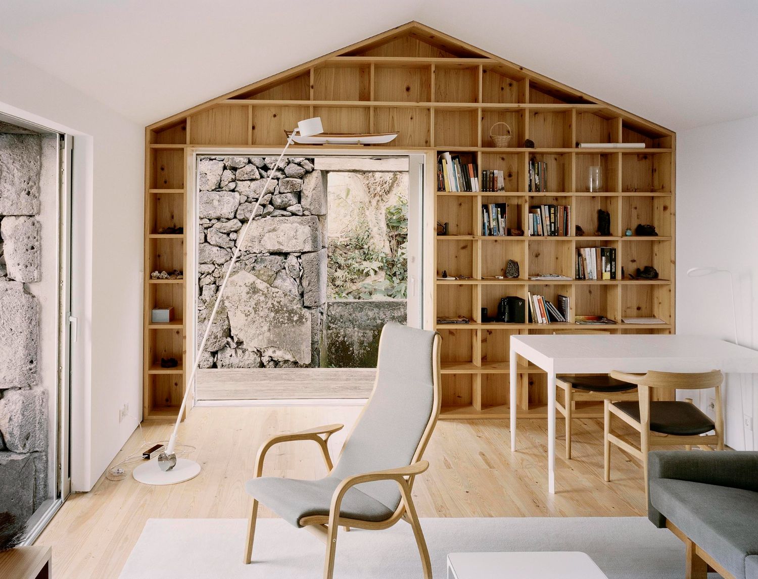E/C House by SAMI-arquitectos — Interior view of the pitched-roof holiday house showing the gable-end wall lined with a timber shelving system of rectangular compartments, stocked with...