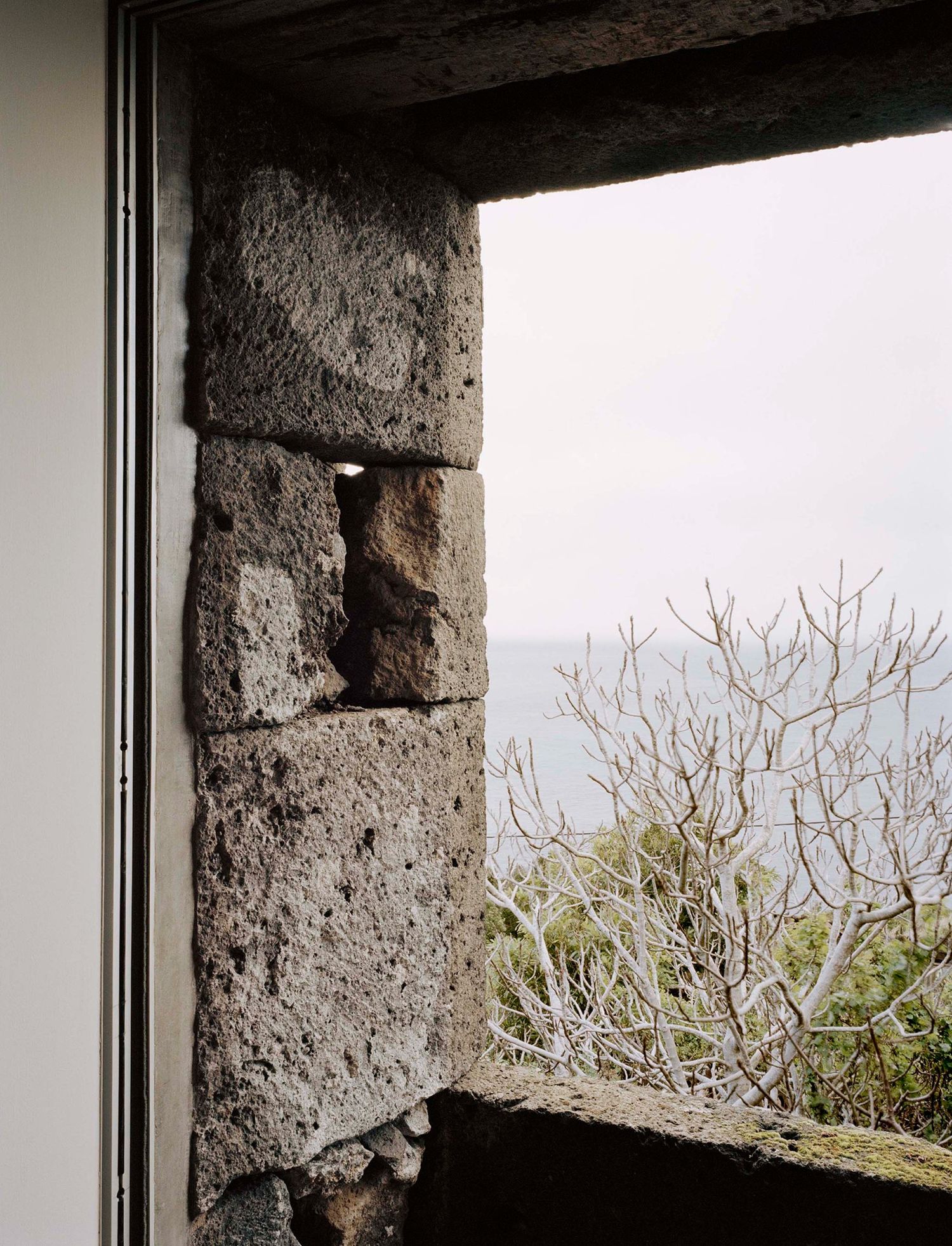 E/C House by SAMI-arquitectos — View through a concrete opening onto weathered stone masonry and landscape beyond.