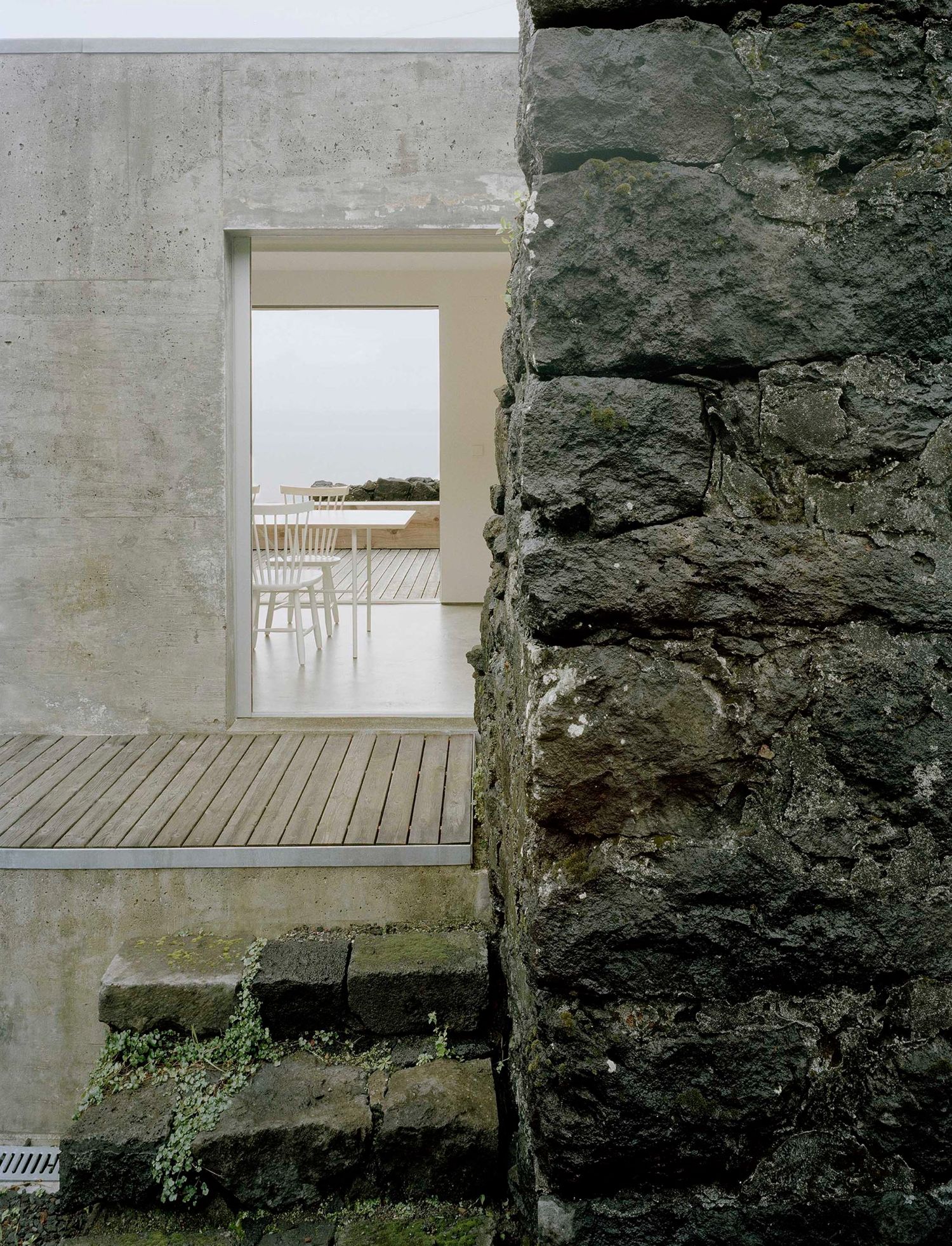 E/C House by SAMI-arquitectos — A weathered volcanic stone wall abuts a board-formed concrete structure, creating a sharp material junction.