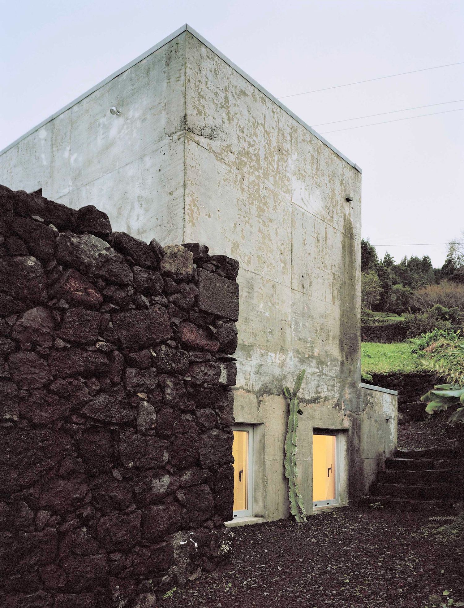 E/C House by SAMI-arquitectos — A concrete structure with a pitched roof and sharp apex sits embedded against a dark volcanic basalt stone wall on Portugal's Island of Pico.