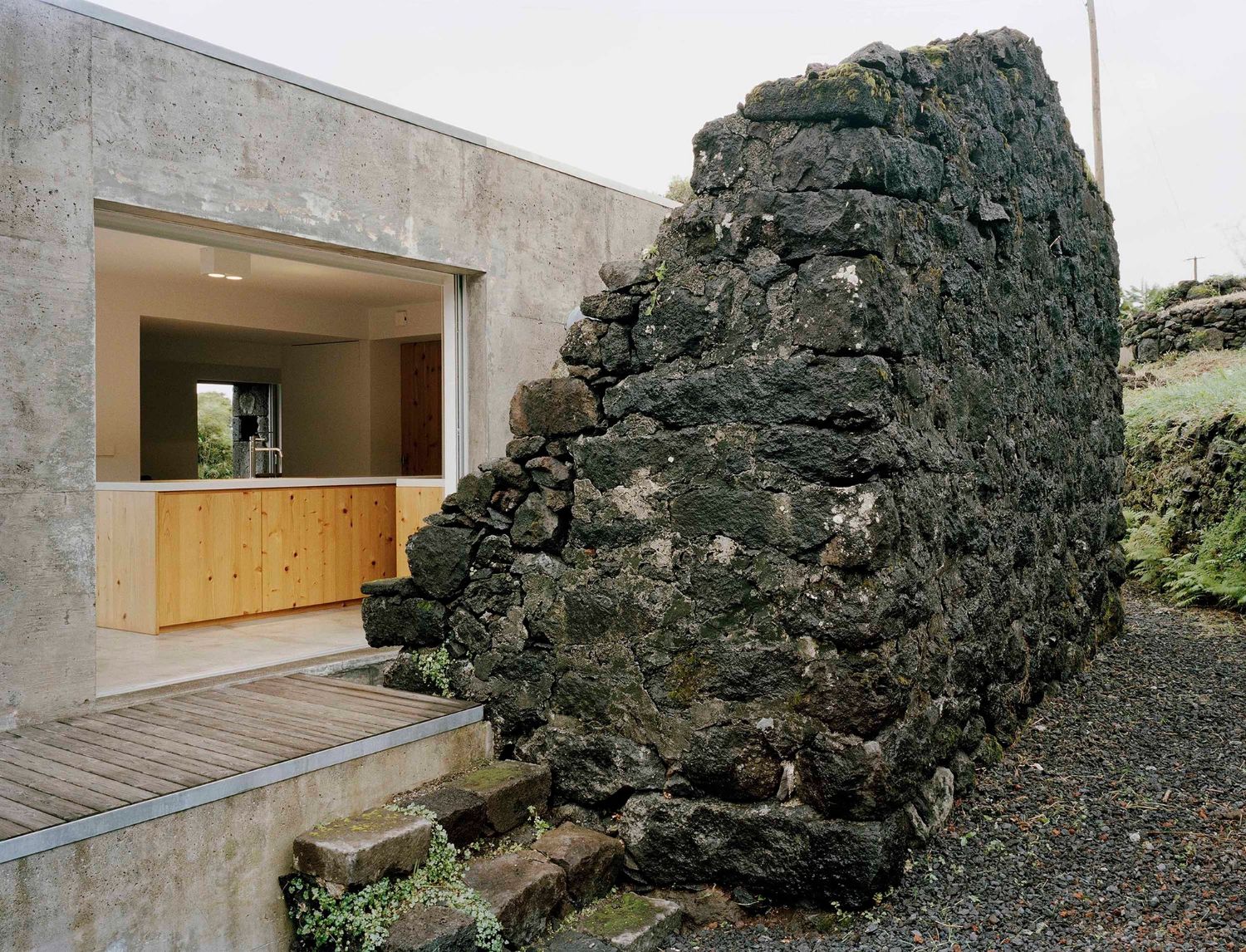 E/C House by SAMI-arquitectos — A modern concrete pavilion with pitched roof abuts a centuries-old volcanic stone wall on Pico Island.