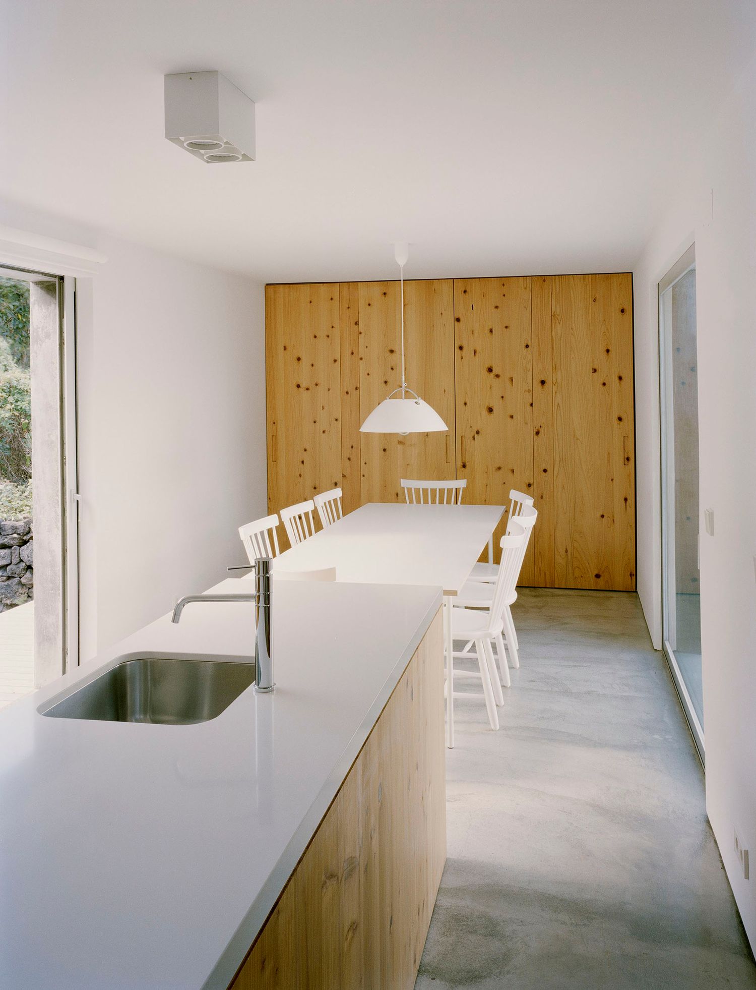 E/C House by SAMI-arquitectos — A linear kitchen-dining space with a white Corian countertop running the length of the left wall, punctuated by a stainless steel sink with chrome faucet.