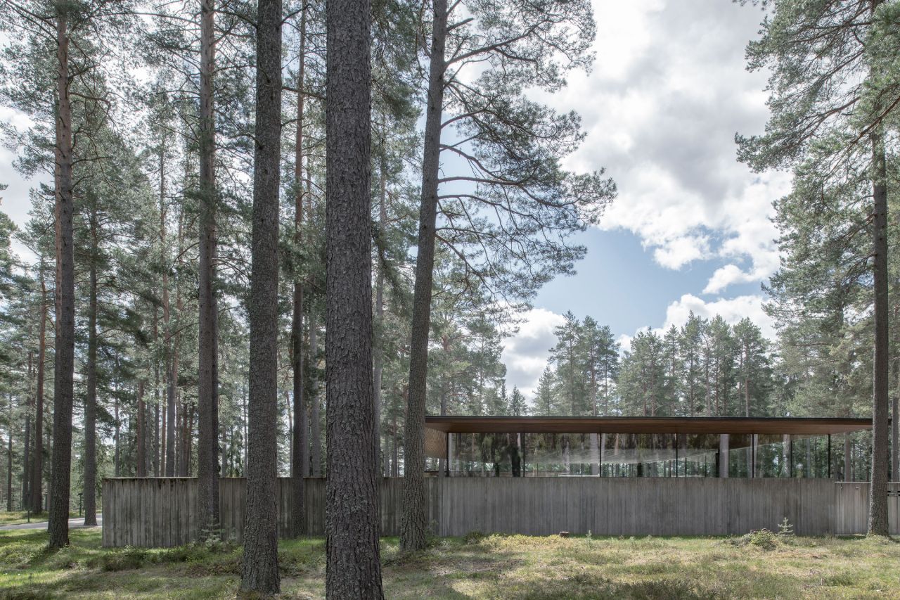 Gävle Crematorium by ELLT captured by Andy Liffner — The Gävle Crematorium's timber-clad long facade integrates seamlessly within a dense coniferous forest, with massive pine trunks punctuating the horizontal...