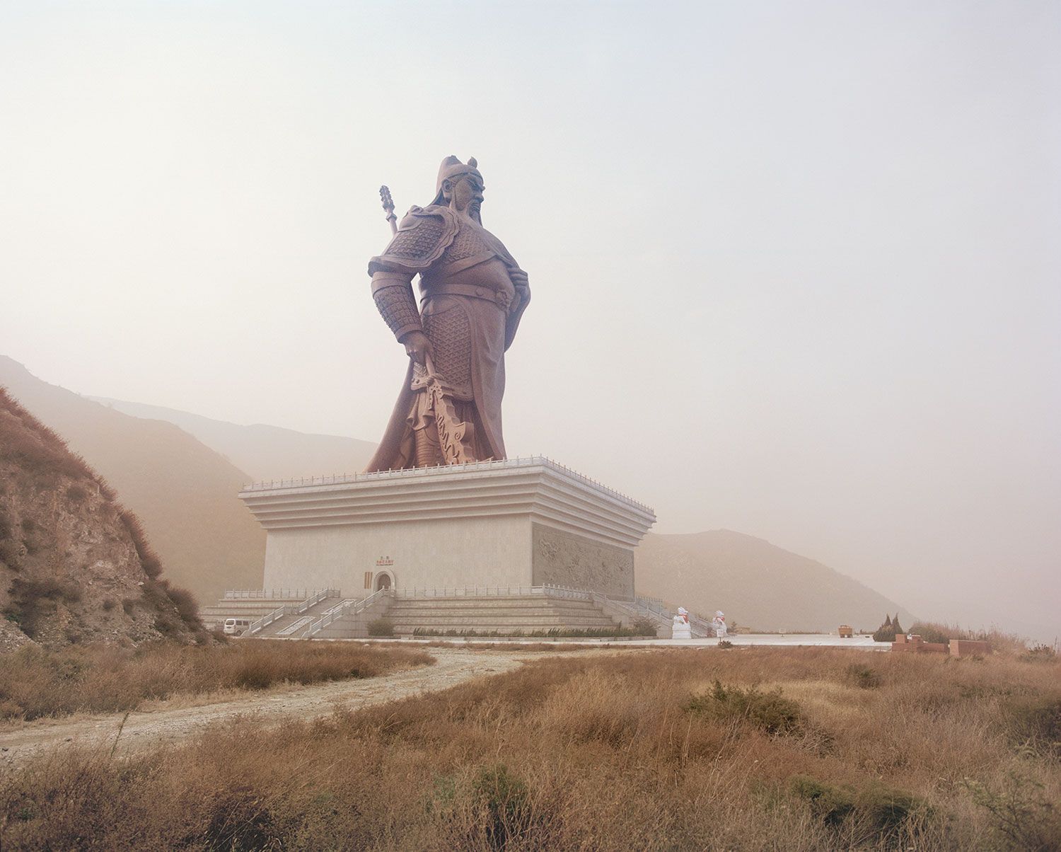 Universal Experience by Catherine Hyland — A bronze warrior monument dominates the composition, rising 57 meters above a tiered concrete base with horizontal striations and metal railings.