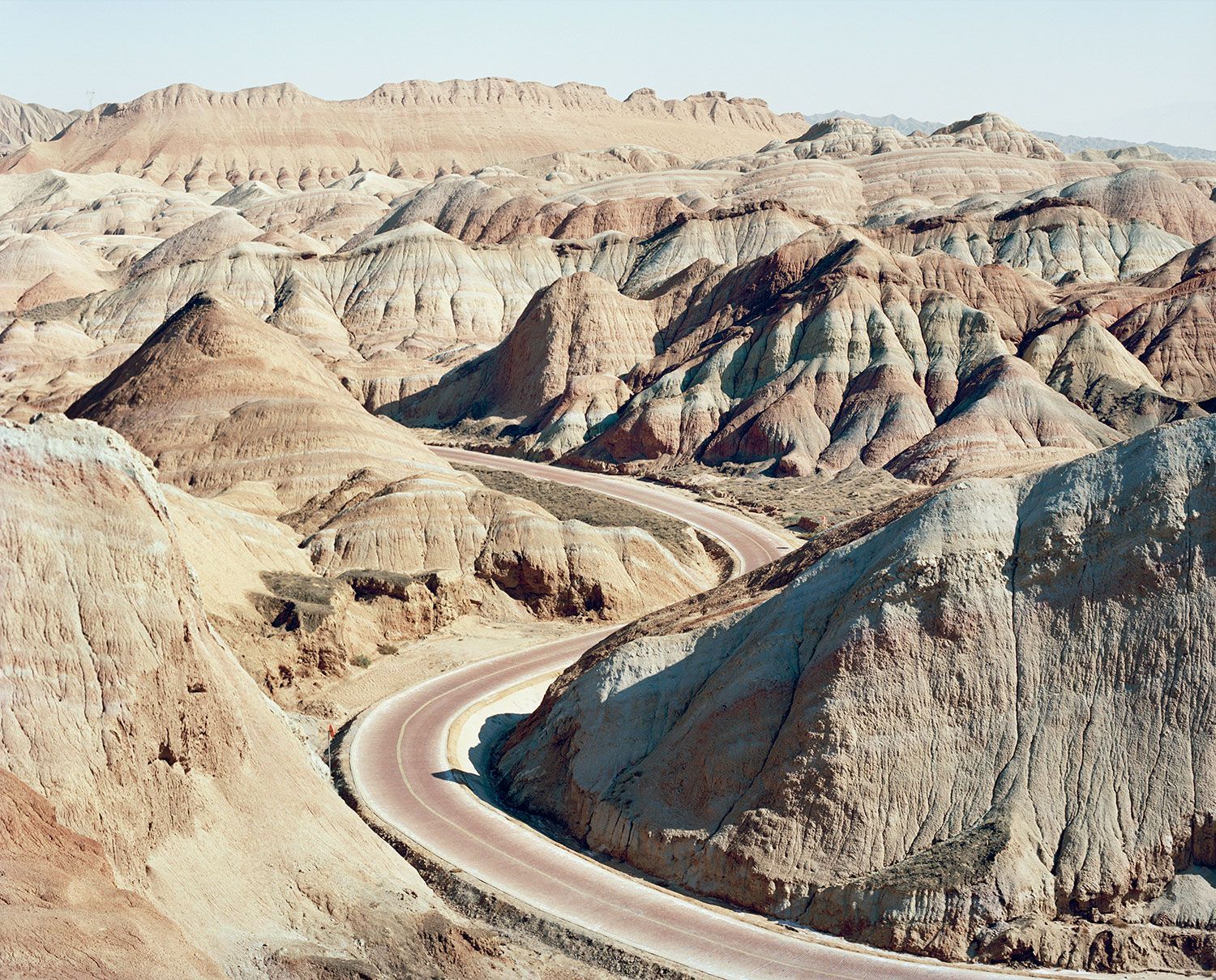 Universal Experience by Catherine Hyland — A winding asphalt road curves sharply through eroded badlands, its pale pink-mauve surface contrasting with surrounding sandstone formations.