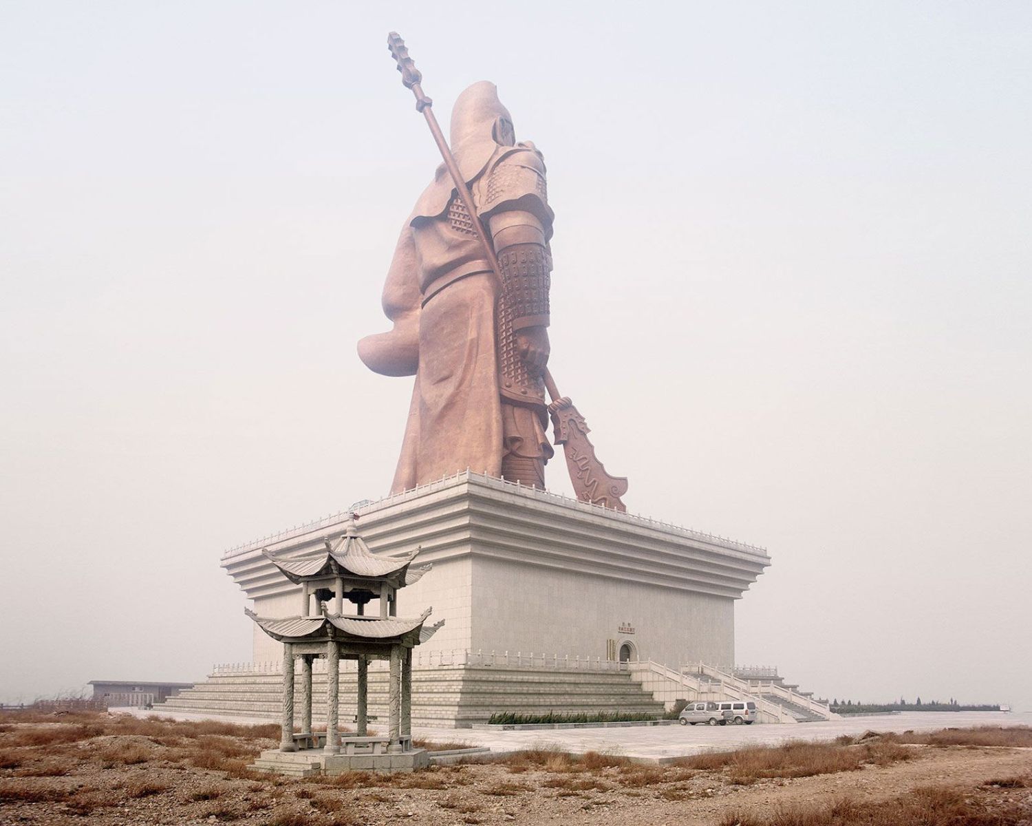 Universal Experience by Catherine Hyland — A 57-meter bronze warrior figure dominates the composition, rendered in oxidized patina with articulated armor plating and studded gauntlets holding an...