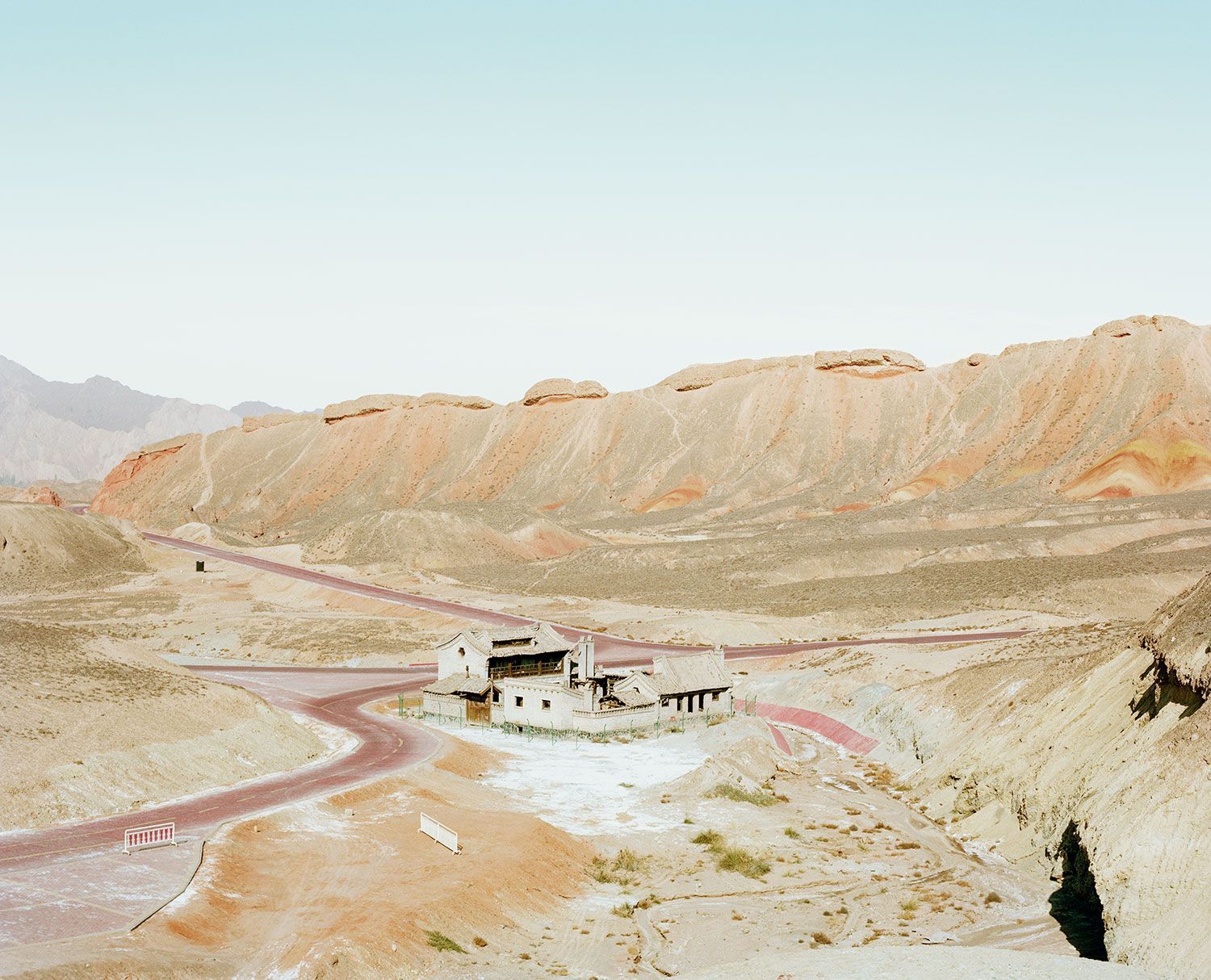 Universal Experience by Catherine Hyland — A modest white-rendered concrete pavilion with dark timber detailing sits isolated in a barren valley, flanked by sinuous rammed-earth roads rendered in...