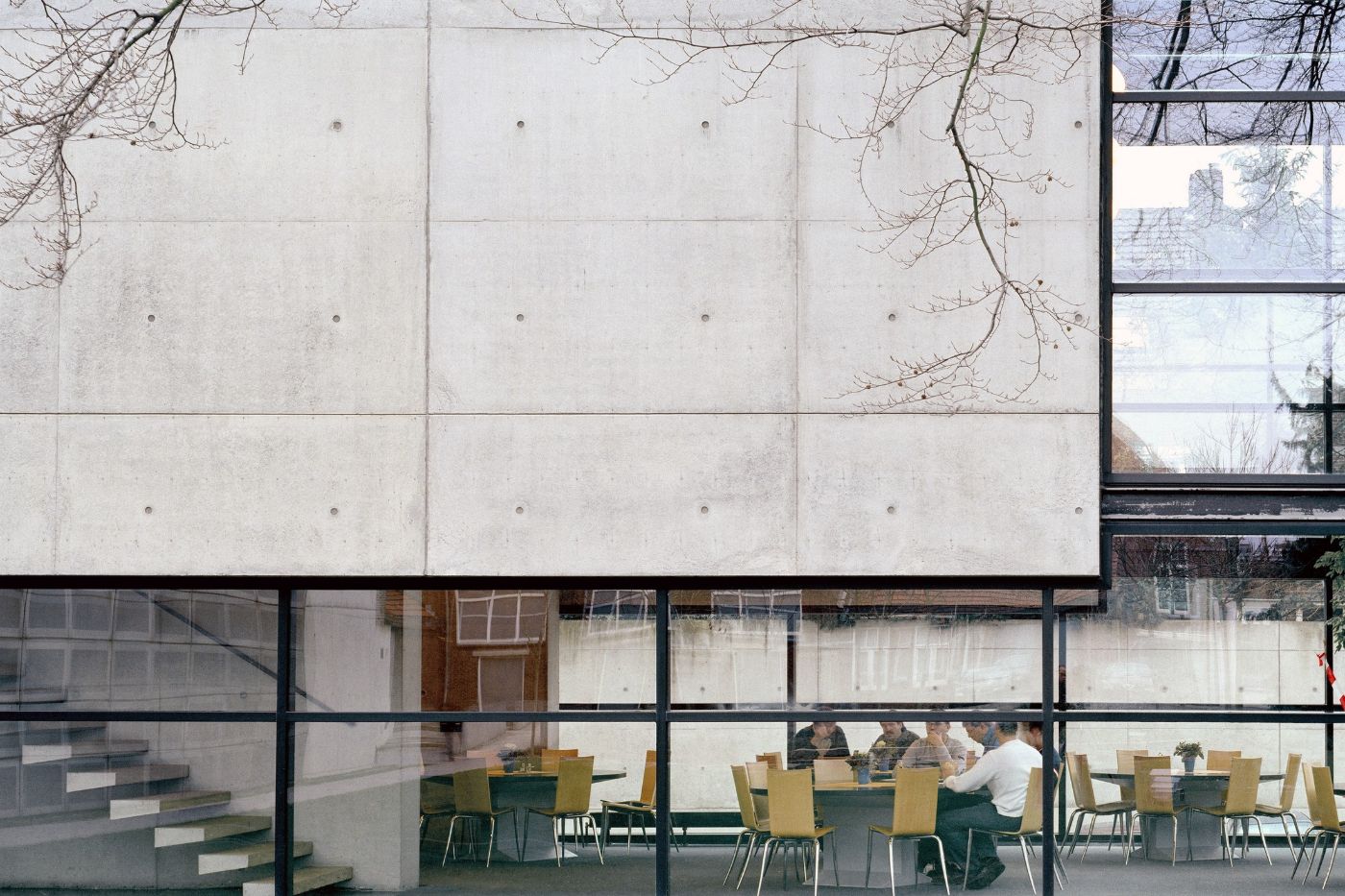 AZL Pension Fund Headquarters by Wiel Arets Architects by WAA Wiel Arets Architects — Ground-level view through continuous black-framed glazing reveals the canteen interior with users seated at tables with yellow-backed chairs.