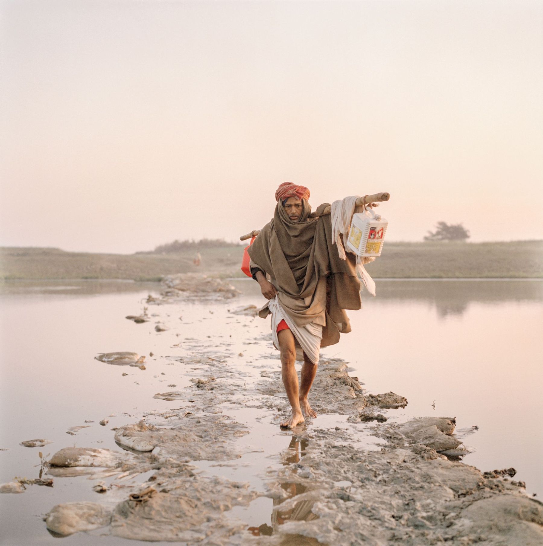Ganga Ma by Giulio Di Sturco — A solitary figure in wrapped earth-toned textiles traverses a rocky causeway through shallow water, pointing toward the distant shoreline.