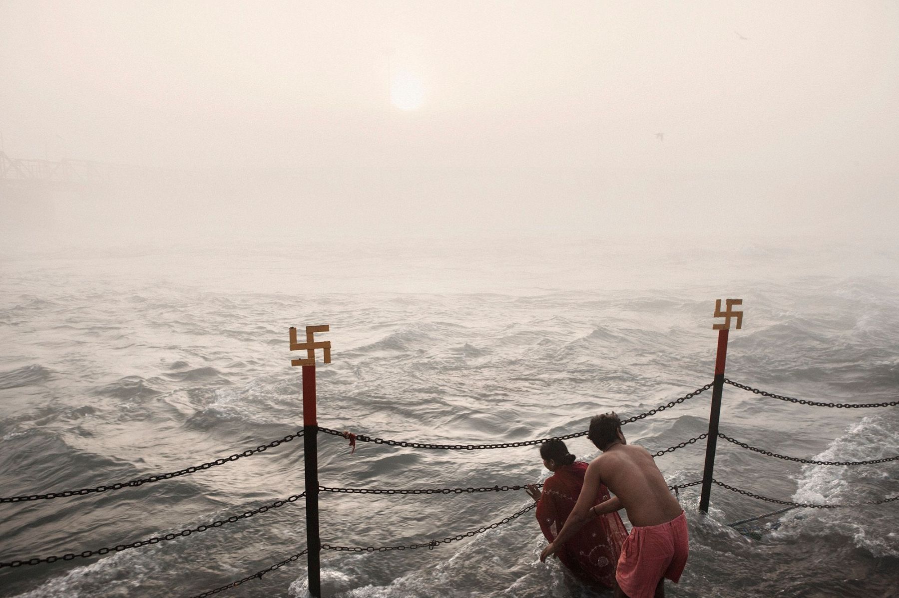 Ganga Ma by Giulio Di Sturco — Two figures stand waist-deep in the Ganges, one wearing a burgundy garment.