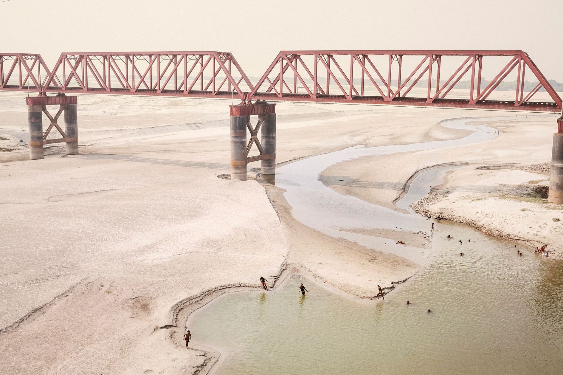 Ganga Ma by Giulio Di Sturco — A steel lattice bridge spans the desiccated riverbed of the Ganges, its geometric framework casting linear shadows across exposed sand and shallow channels...