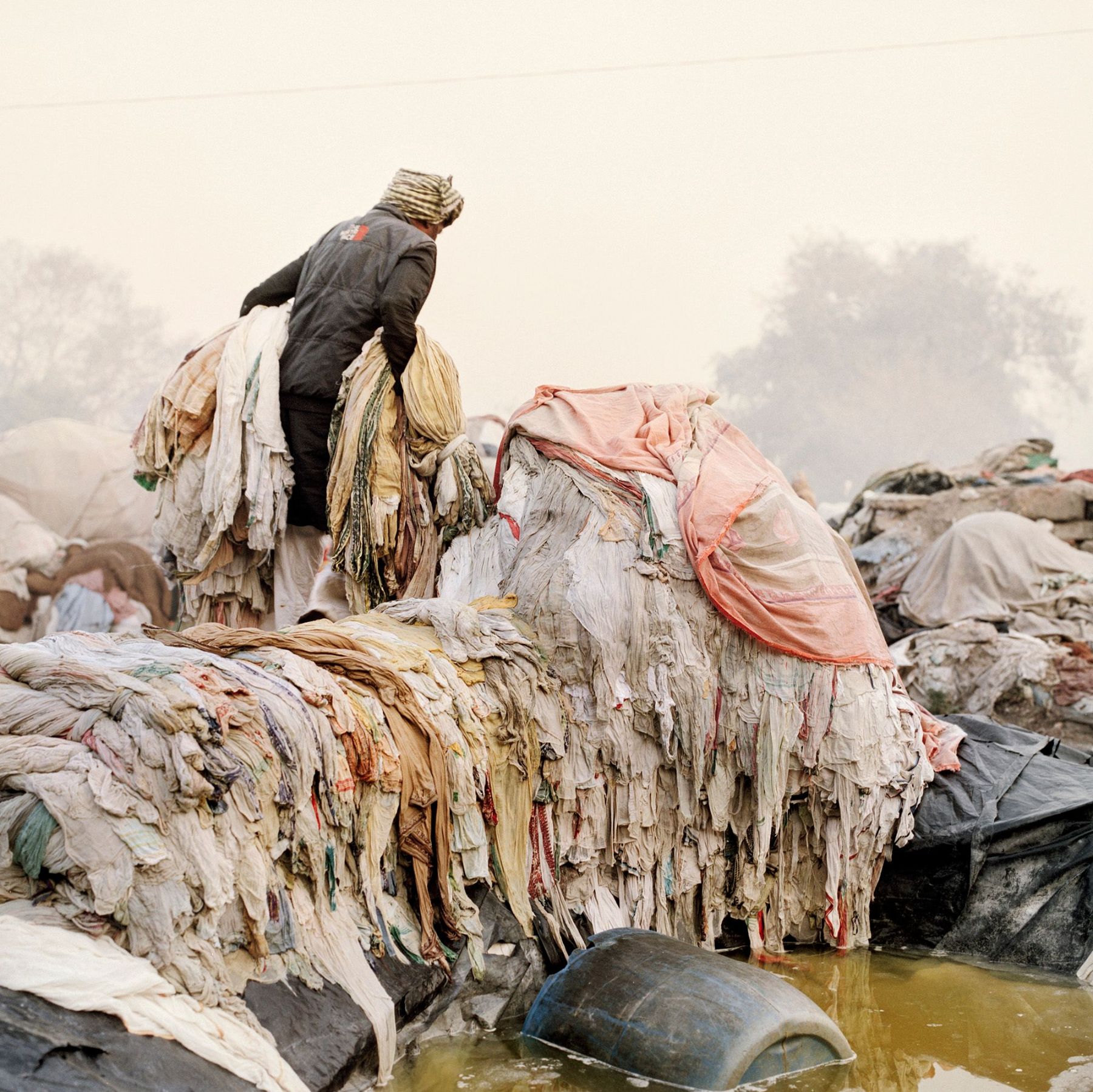 Ganga Ma by Giulio Di Sturco — A worker stands atop compressed bundles of textiles and refuse at the river's edge, their figure silhouetted against the hazy landscape.