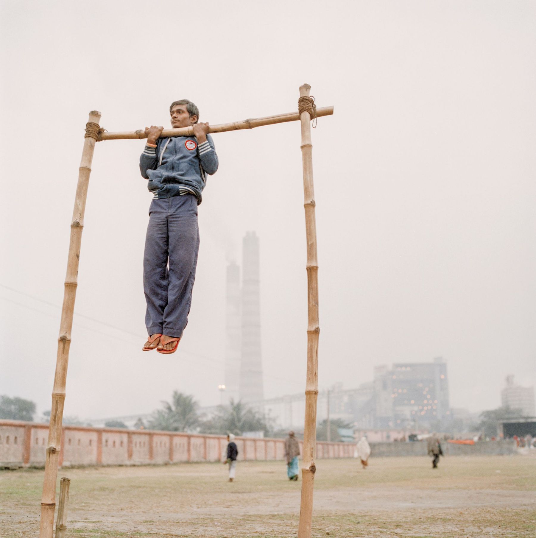 Ganga Ma by Giulio Di Sturco — A young man performs a chin-up on a bamboo bar suspended between two vertical bamboo poles in an open field.