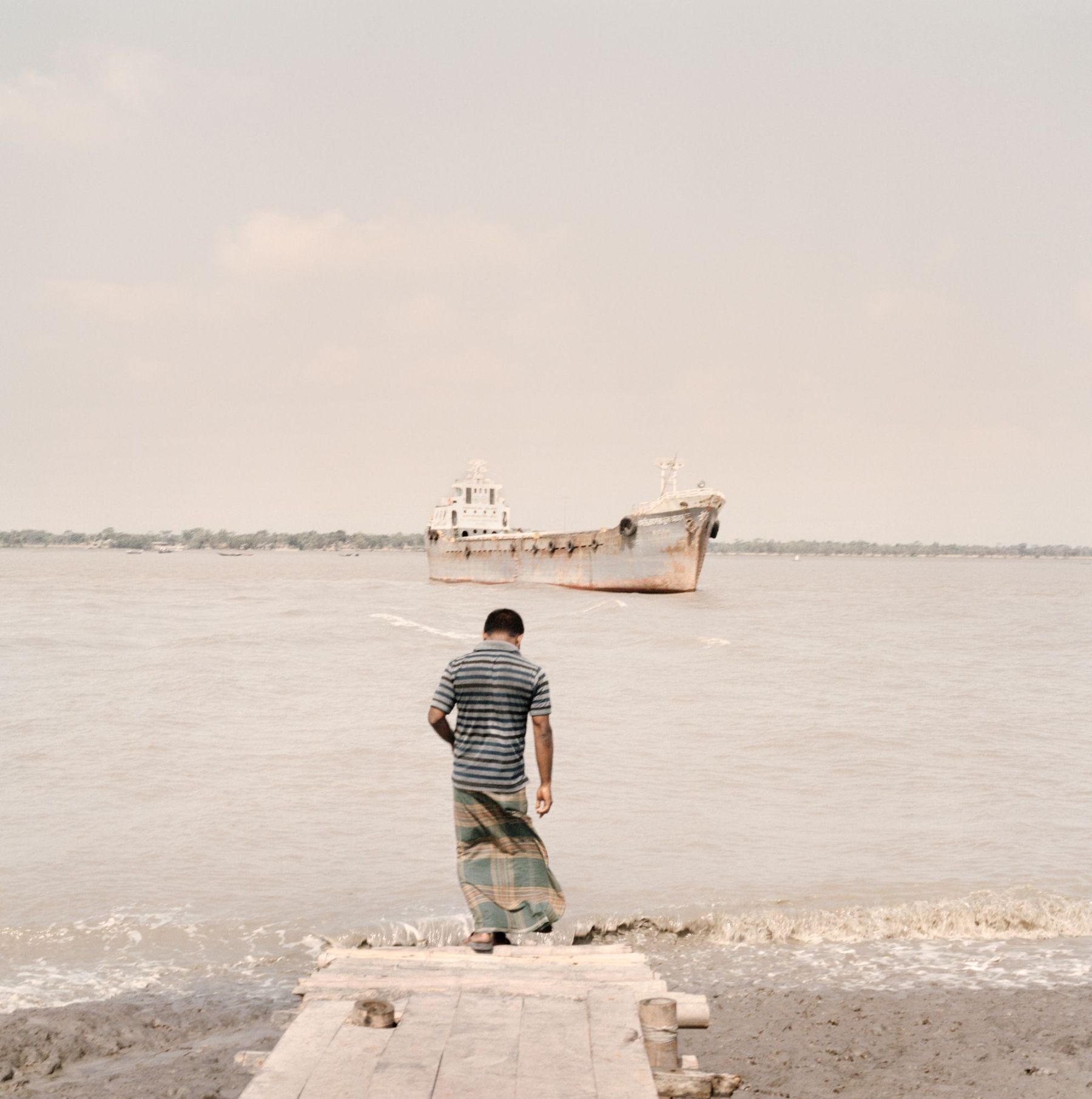 Ganga Ma by Giulio Di Sturco — A solitary figure in striped shirt stands on a concrete platform extending into shallow water, facing an abandoned cargo vessel with visible rust patination...
