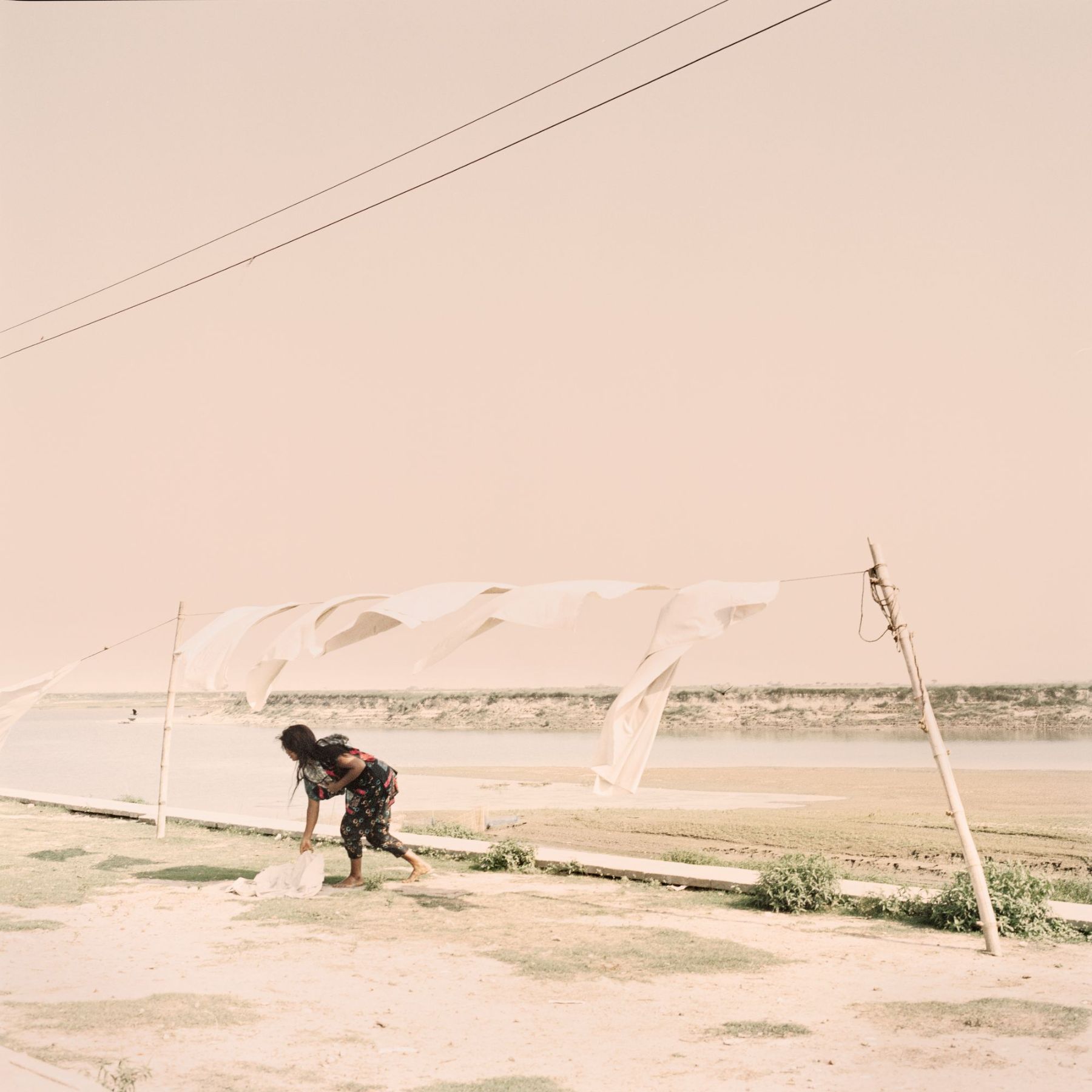 Ganga Ma by Giulio Di Sturco — A solitary figure bends forward in shallow water, gathering bundled dried vegetation along the riverbank.