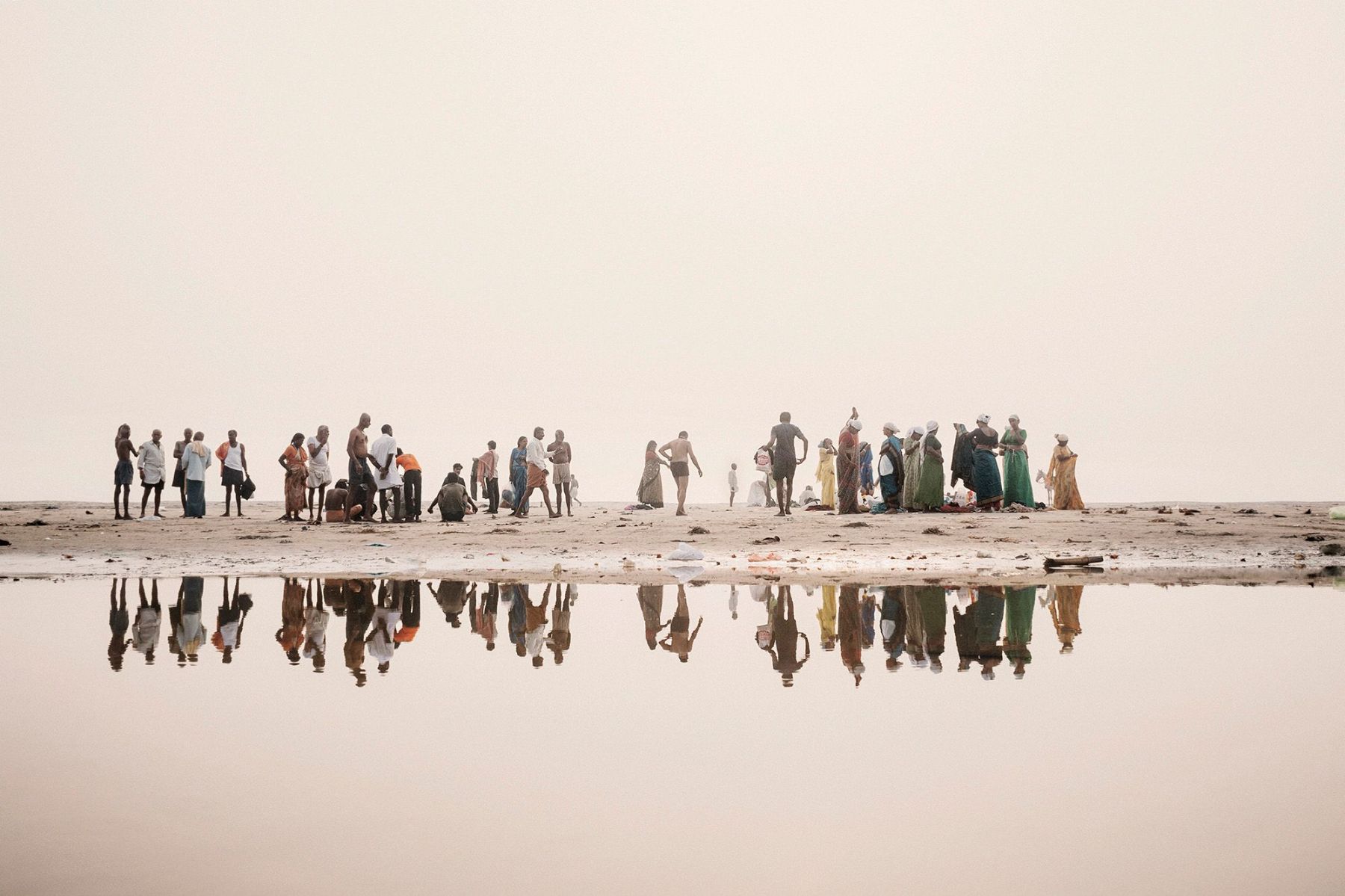 Ganga Ma by Giulio Di Sturco — A linear gathering of approximately forty figures stands along the exposed mudflat of the Ganges, their reflections mirrored in shallow water that divides...