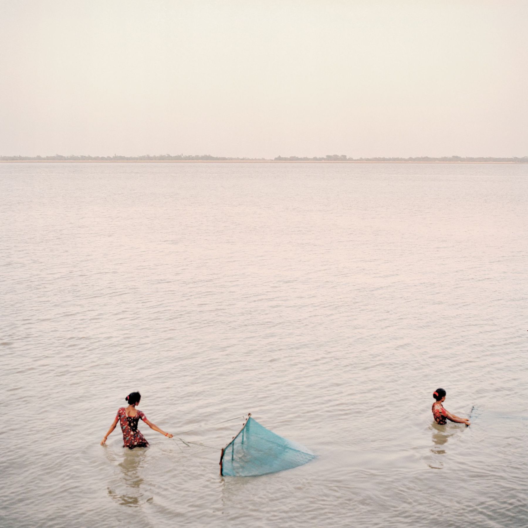 Ganga Ma by Giulio Di Sturco — Two figures in patterned cotton garments wade in shallow water, deploying a turquoise fishing net in the center frame.