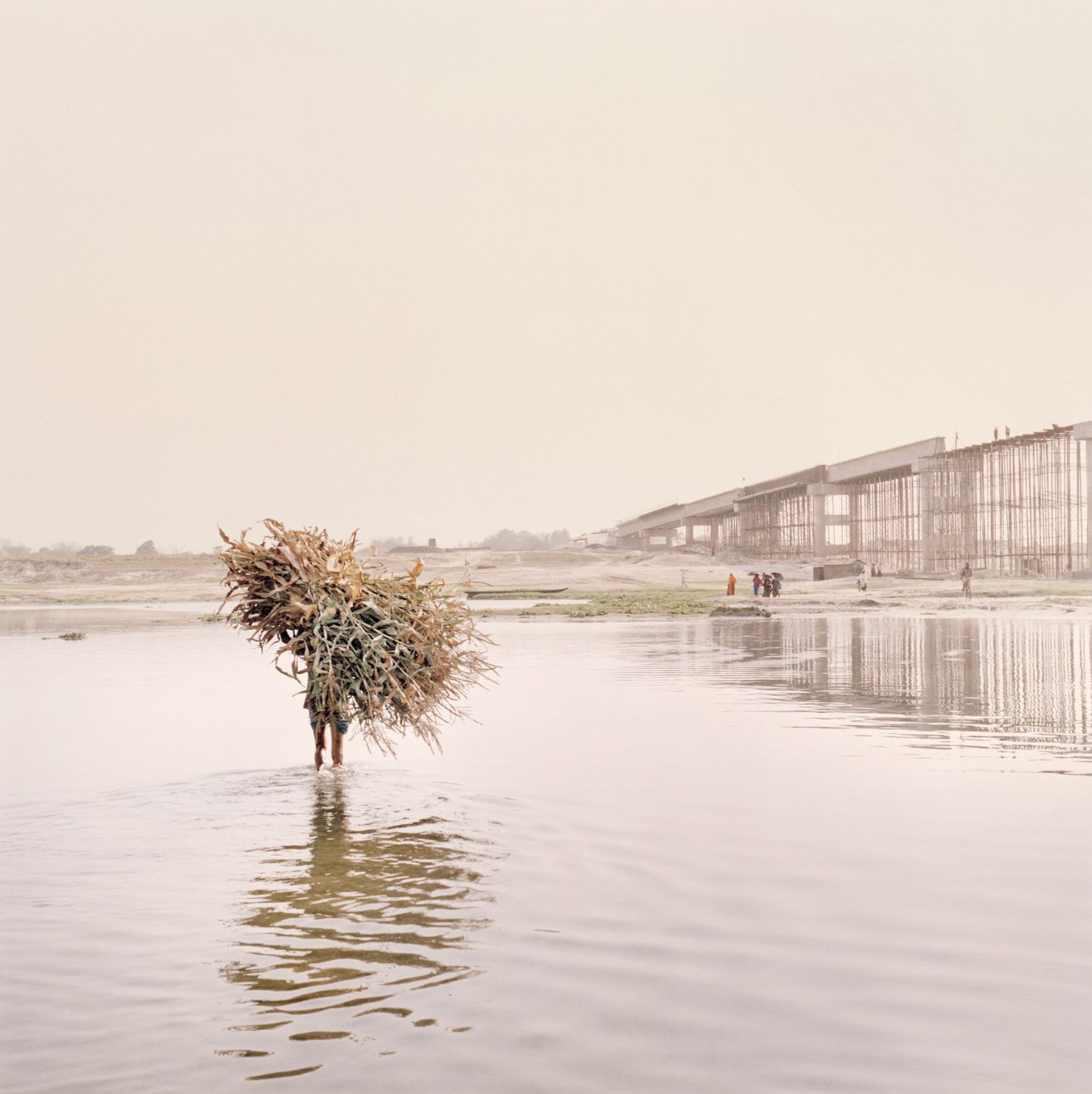 Ganga Ma by Giulio Di Sturco — A solitary figure wades through shallow water carrying a large bundle of dried vegetation and straw, their silhouette stark against the expansive floodplain.