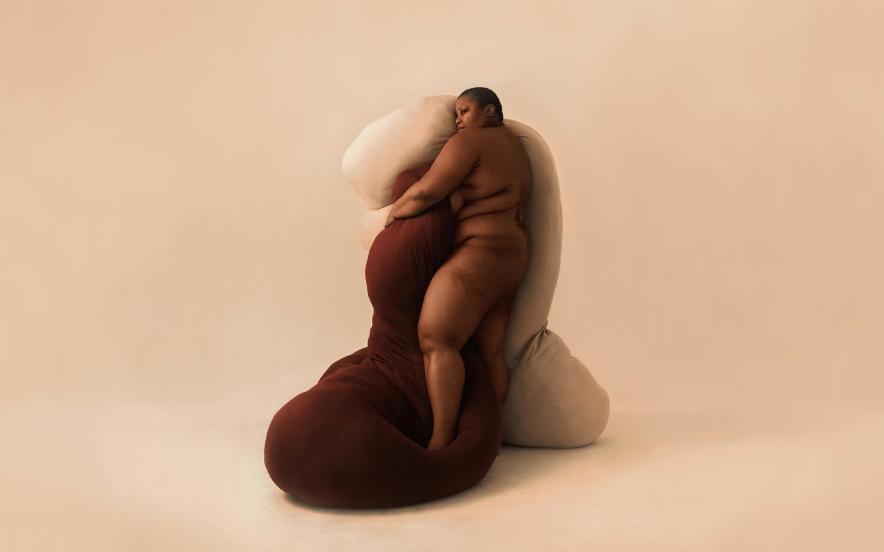 Selected works by Lakin Ogunbanwo — A studio photograph featuring two human figures in close physical contact positioned against a large cream-colored soft sculptural form with organic,...