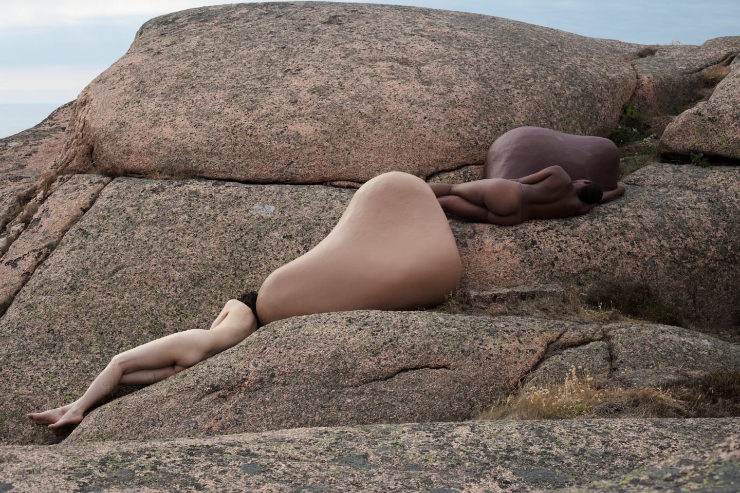 Selected works by Lakin Ogunbanwo — Two nude figures positioned across a large granite boulder formation in an outdoor landscape setting.