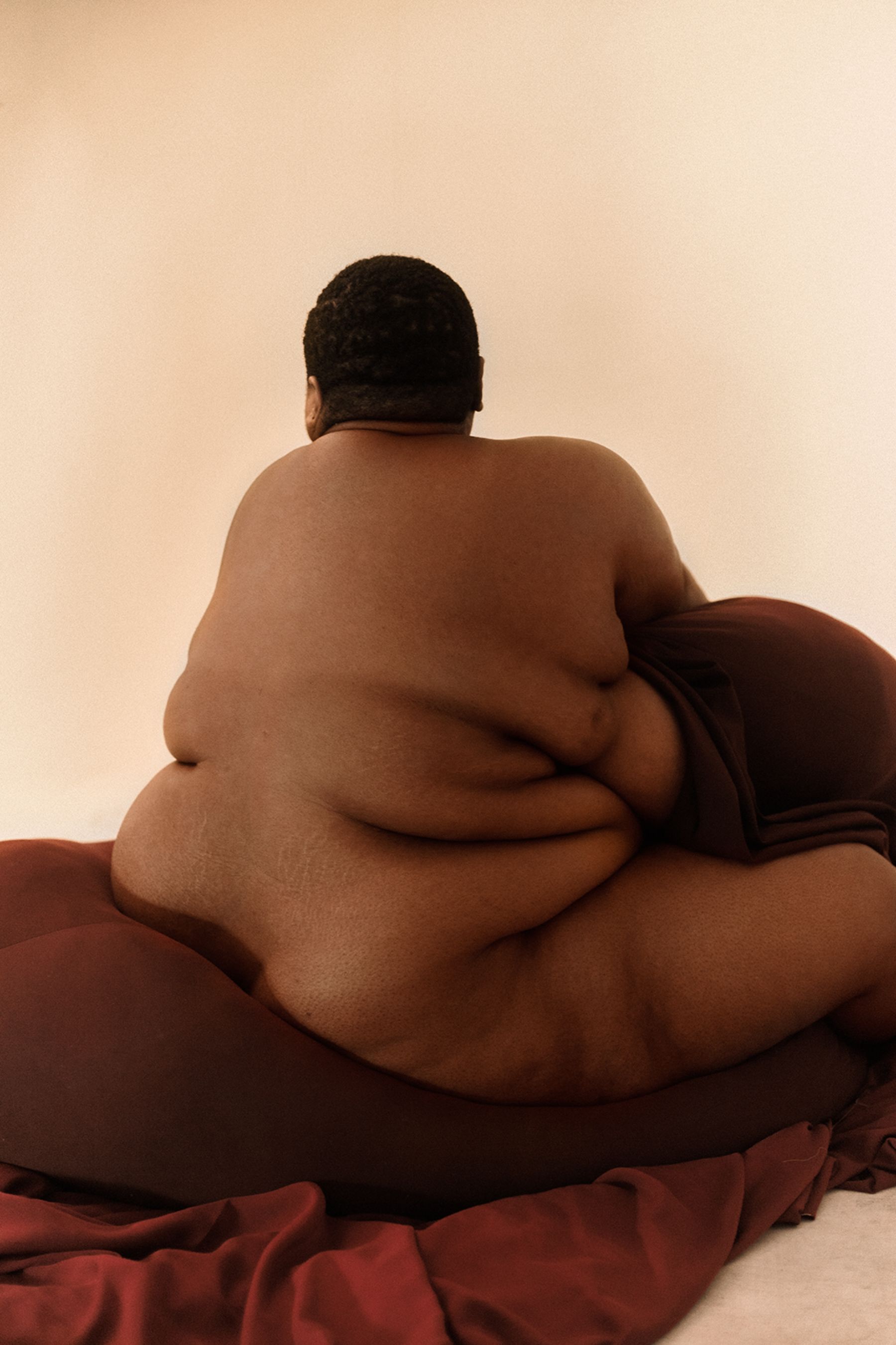 Selected works by Lakin Ogunbanwo — A seated figure with substantial volumetric form dominates the composition, back to camera against a neutral beige ground.
