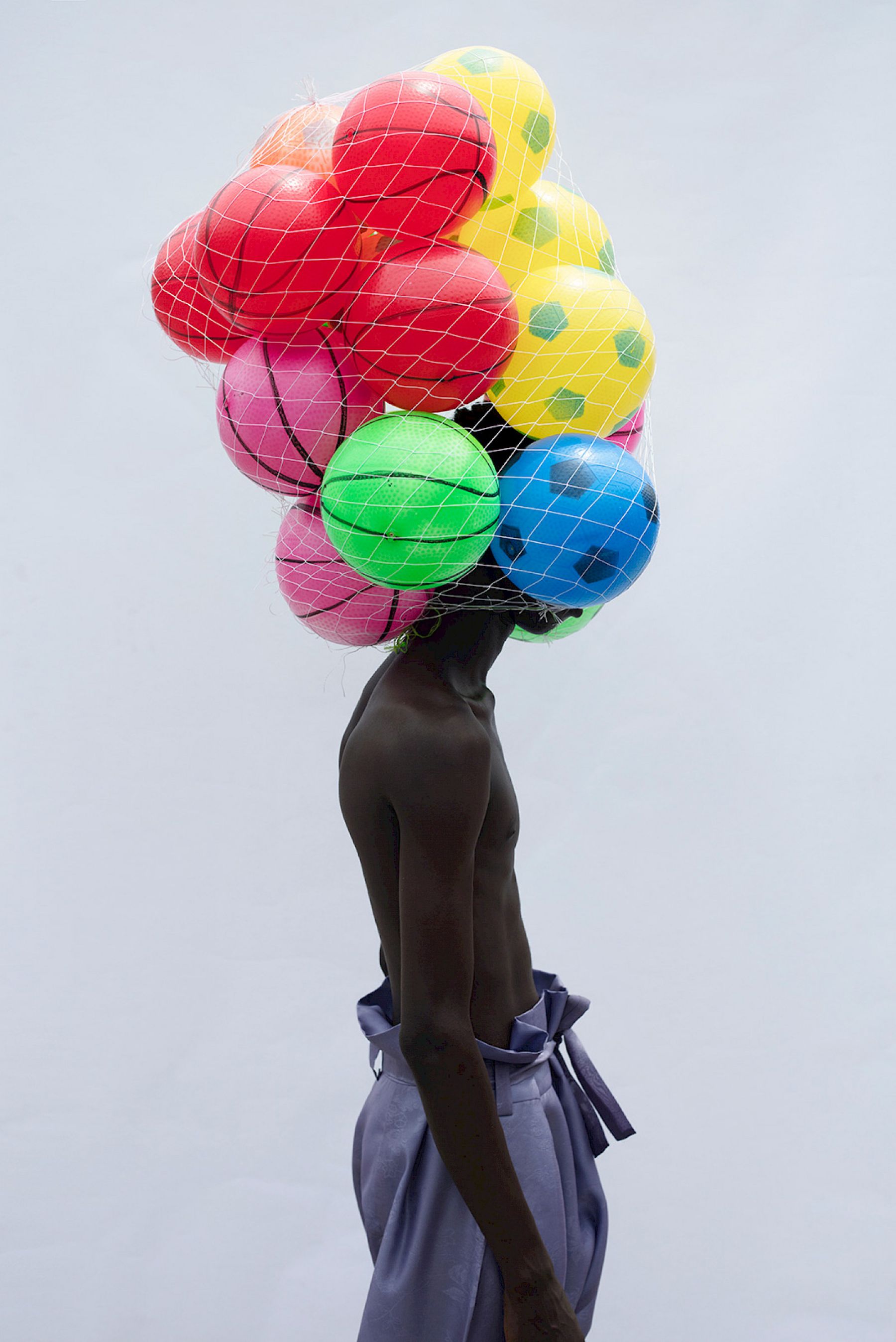 Selected works by Lakin Ogunbanwo — A figure stands against a neutral ground, torso obscured by a mesh net containing inflatable balls in vibrant primaries—red, yellow, blue, green—and...