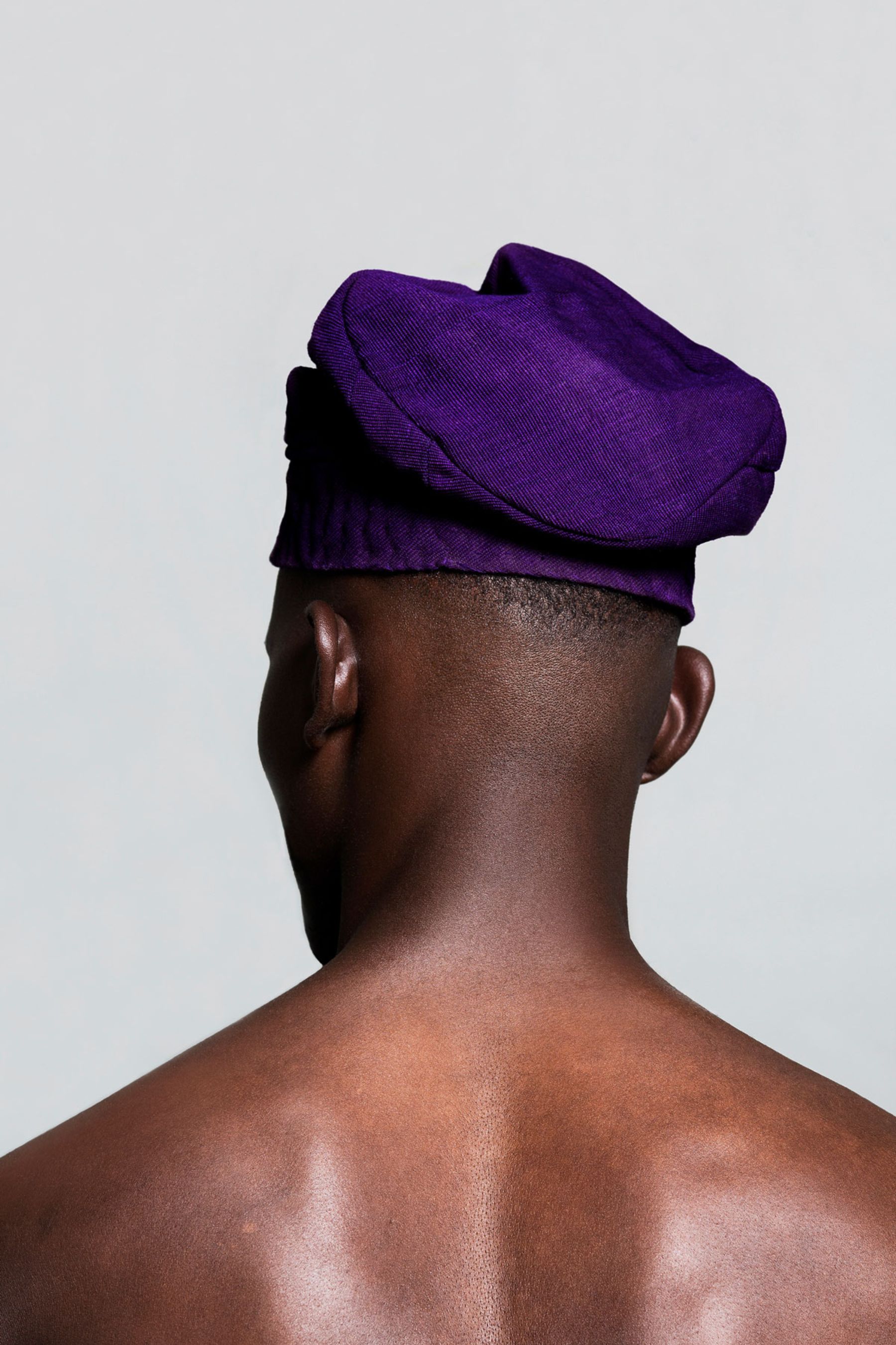 Selected works by Lakin Ogunbanwo — Rear portrait of a shirtless male figure wearing a deep purple ribbed cap tilted at an angle, shot against a neutral cream background.