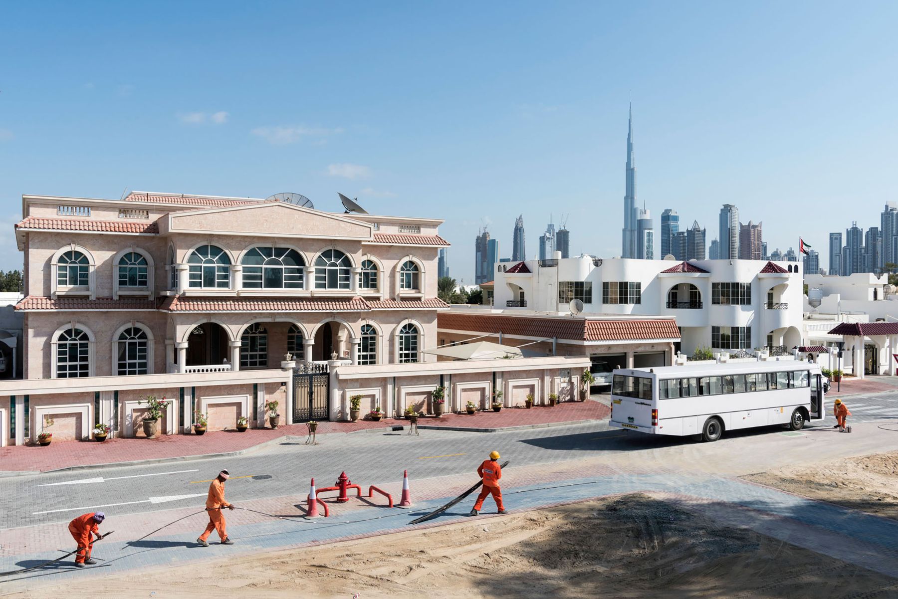 Garden of Delight by Nick Hannes — Construction workers in safety orange lay asphalt on a plaza fronting a palatial beachfront complex in Dubai.