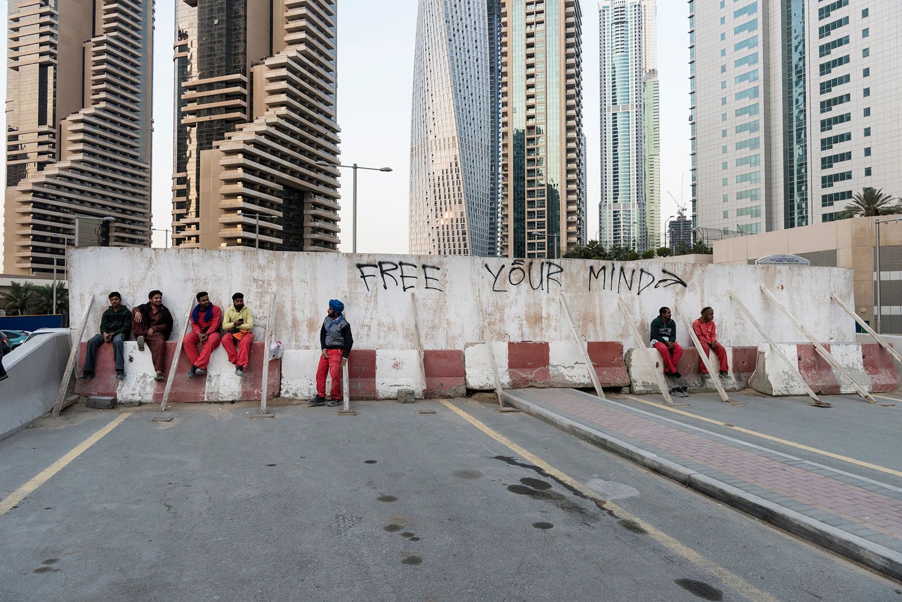 Garden of Delight by Nick Hannes — A concrete barrier wall inscribed with 'FREE YOUR MIND' dominates the foreground of this Dubai parking structure, where eight figures in coordinated red and...