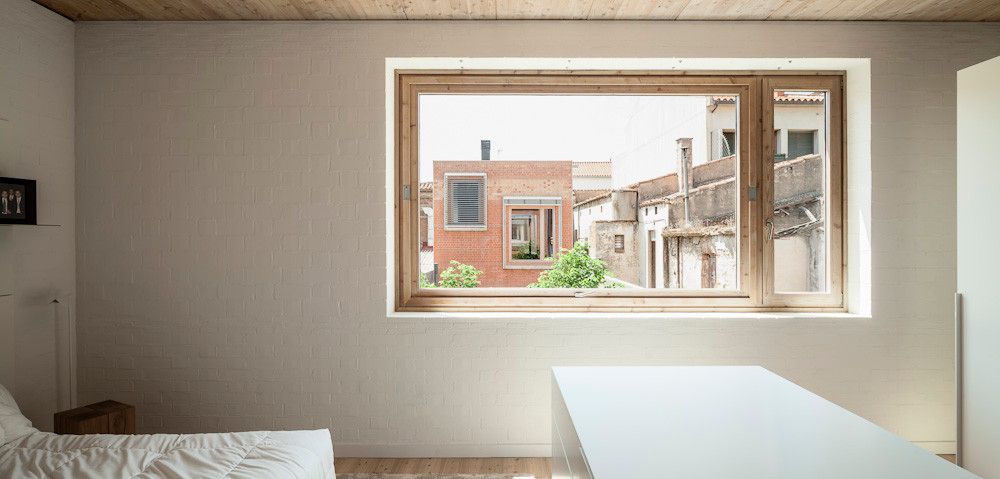 House 1014 by H Arquitectes — A bedroom interior framed by a large timber-clad window overlooking the narrow urban streetscape of Granollers' historical centre.