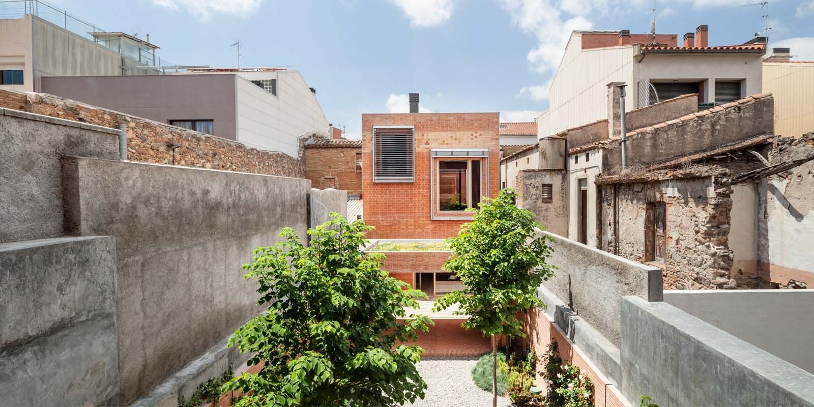 House 1014 by H Arquitectes — A narrow courtyard garden accessed from street level, framed by flanking concrete party walls with weathered patina.