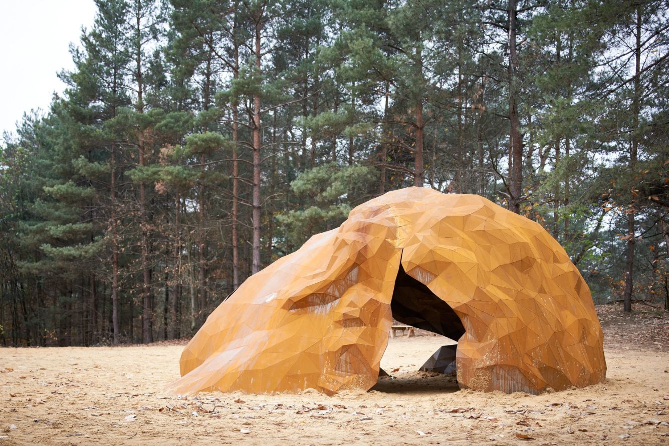 A Giant Sculpture by Gijs Van Vaerenbergh — A monumental welded-steel sculpture with oxidized corten patina occupies a sunken sandy clearing in a pine forest.