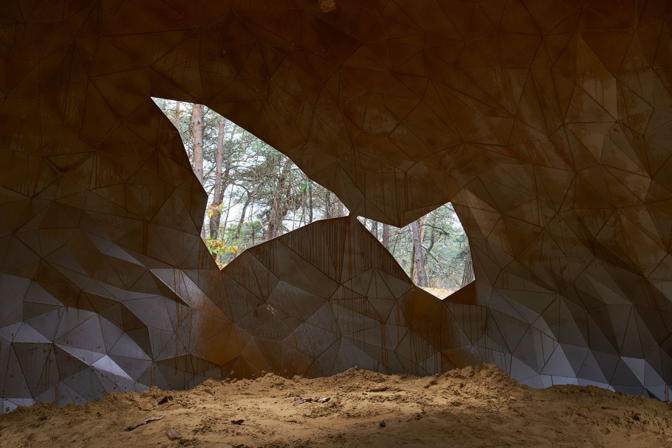 A Giant Sculpture by Gijs Van Vaerenbergh — A monumental steel sculpture with oxidized corten surface dominates a sunken sandy clearing, its faceted geometric planes forming an abstracted head with...