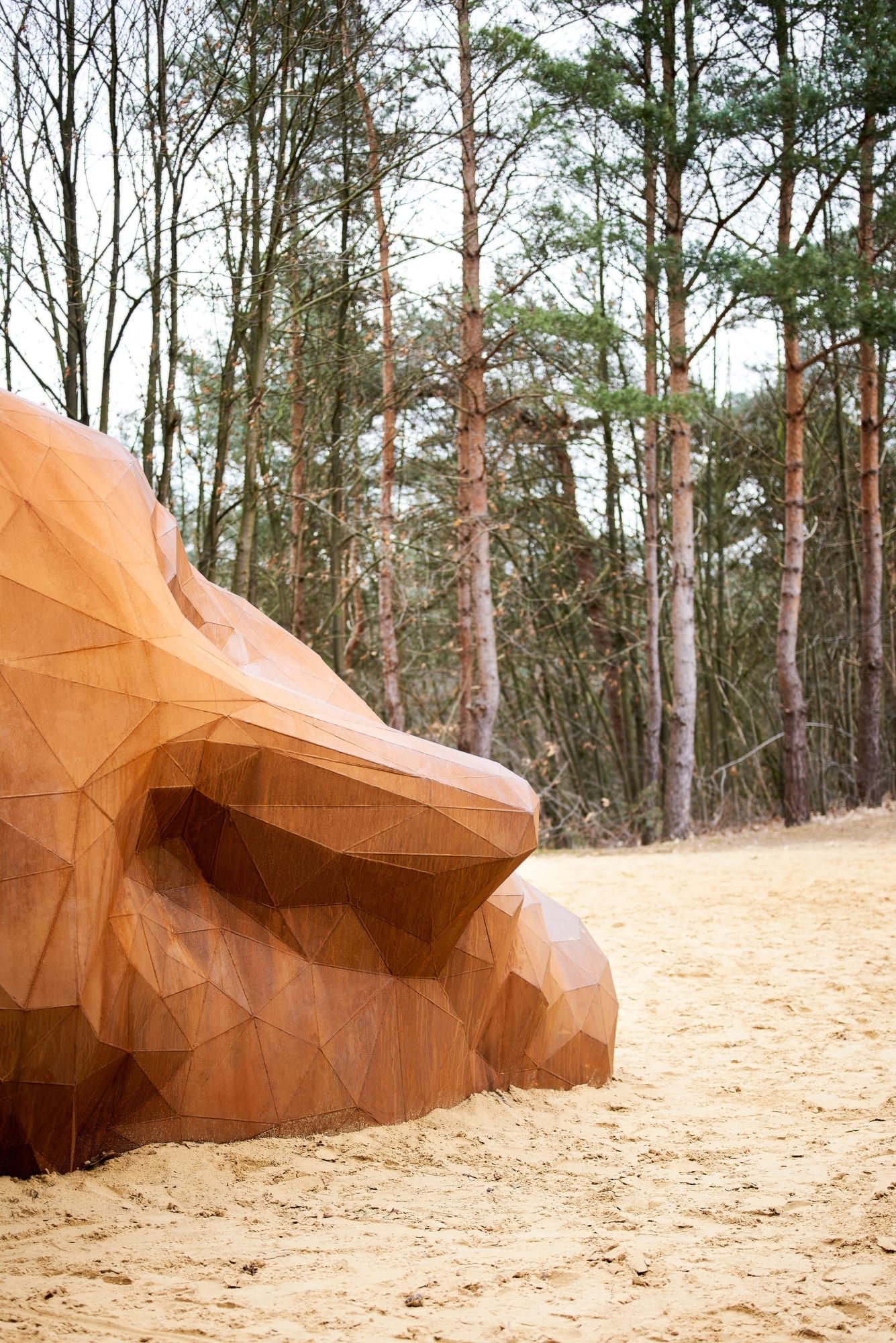 A Giant Sculpture by Gijs Van Vaerenbergh — A monumental wooden sculpture carved in faceted geometric planes occupies a sunken sandy clearing in Kasterlee, Belgium.